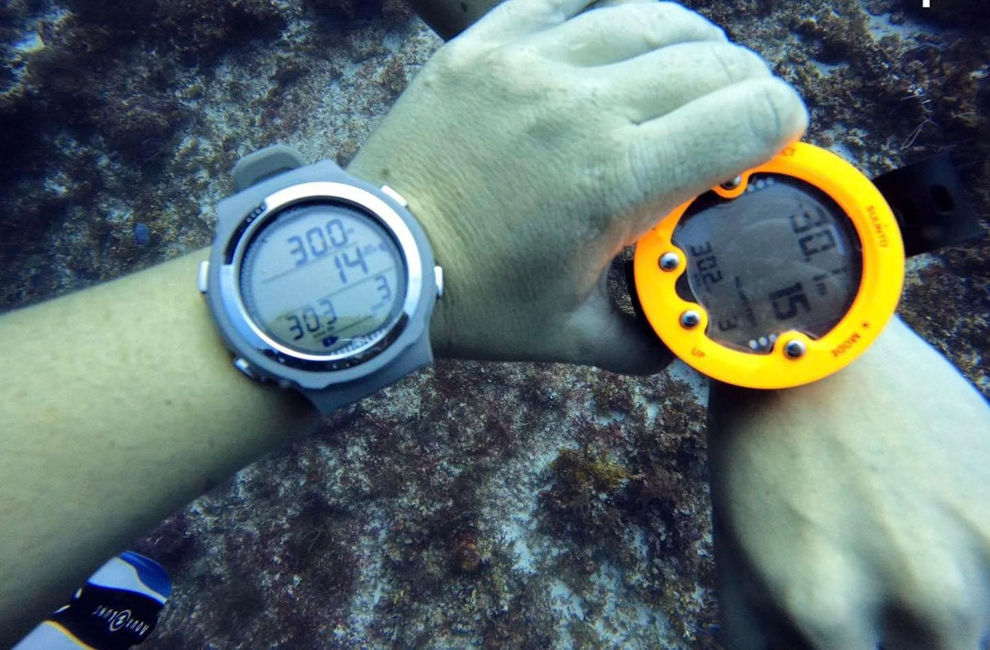 Person underwater wearing a yellow glove and a grey diving watch, checking a yellow digital dive computer, with rocky ocean floor in the background.  Cozumel Scuba Diving Dive with k8