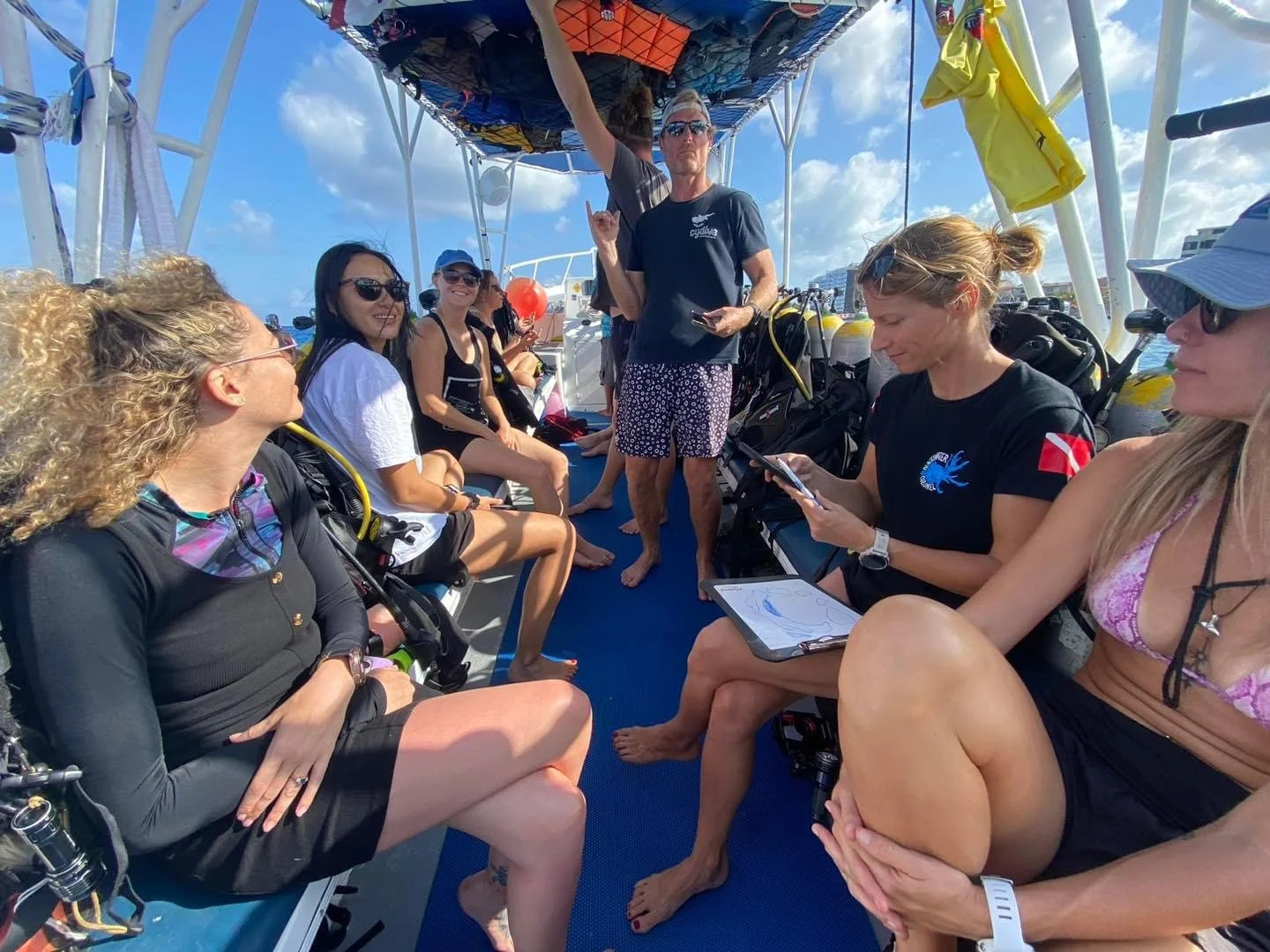 Group of people on a boat, some in scuba gear, with diving equipment, under a blue sky with clouds, preparing for a dive or explaining plans.  Cozumel Scuba Diving Dive with k8