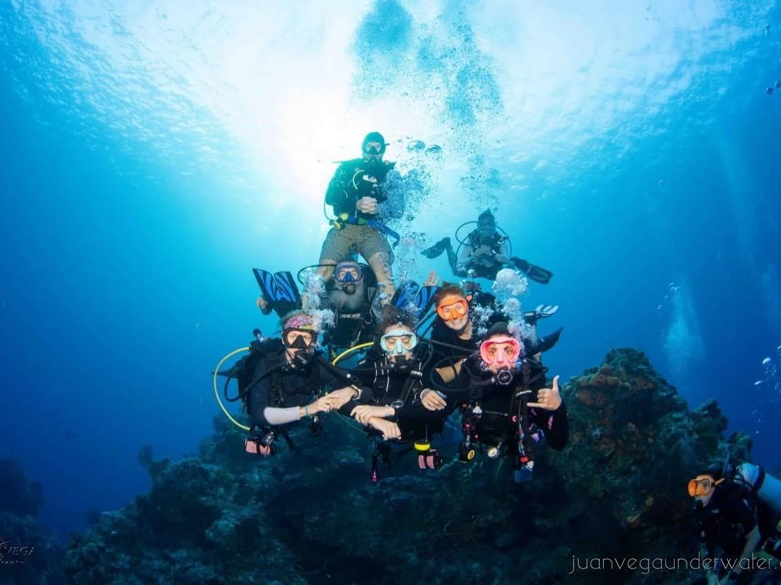 Group of six scuba divers underwater near coral reef, wearing masks and wetsuits, some giving hand signals.  Cozumel Scuba Diving Dive with k8