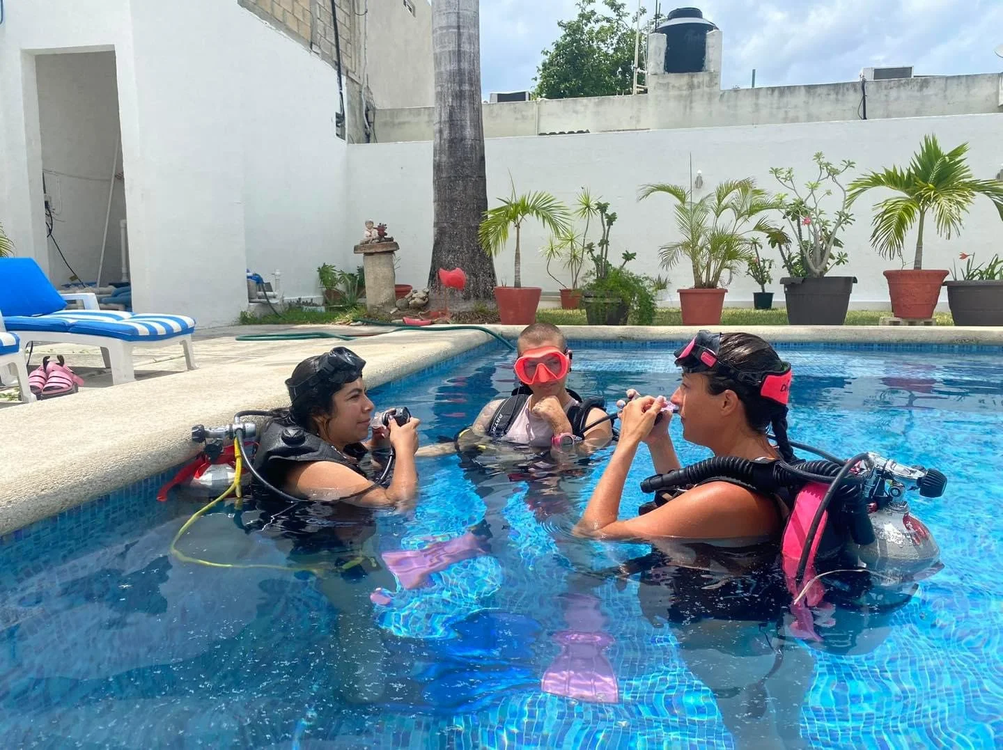 Three women in scuba diving gear standing in a swimming pool, talking, with poolside chairs and potted plants in the background.  Cozumel Scuba Diving Dive with k8