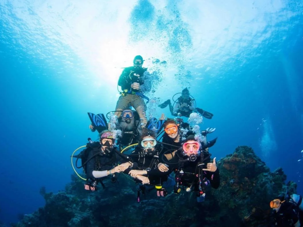 Group of six scuba divers underwater, some making gestures, surrounded by blue water and coral.  Cozumel Scuba Diving Dive with k8