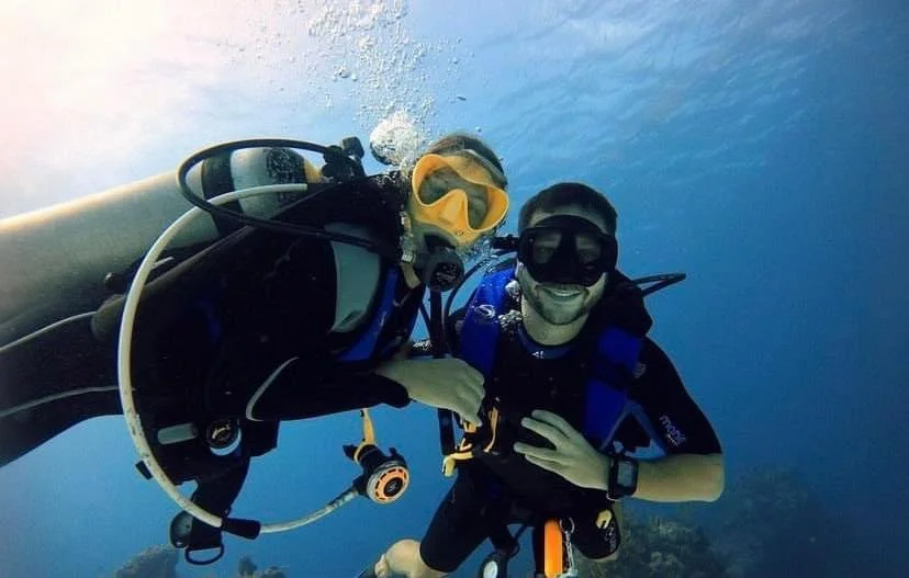 Two scuba divers underwater, smiling and taking a selfie, with clear blue water in the background.  Cozumel Scuba Diving Dive with k8