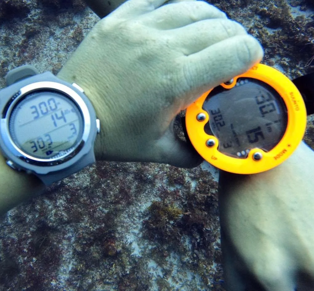 Close-up of two diver's wrists showing digital watches with underwater backgrounds; one watch is gray with a white face, the other is orange with a gray face.  Cozumel Scuba Diving Dive with k8