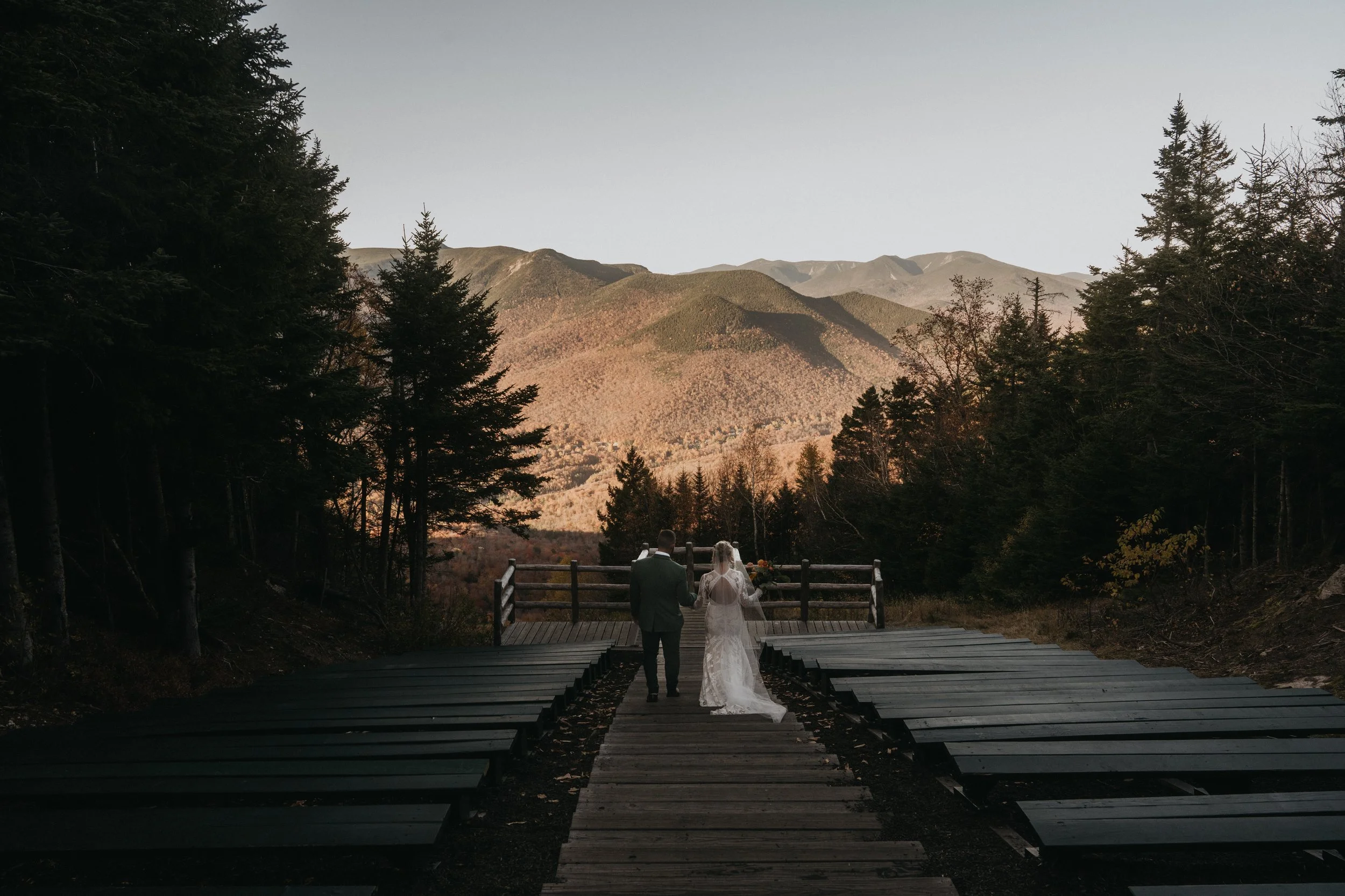 A bride and groom walk hand in hand on a wooden path in a forested mountain landscape during sunset, with mountains in the background.