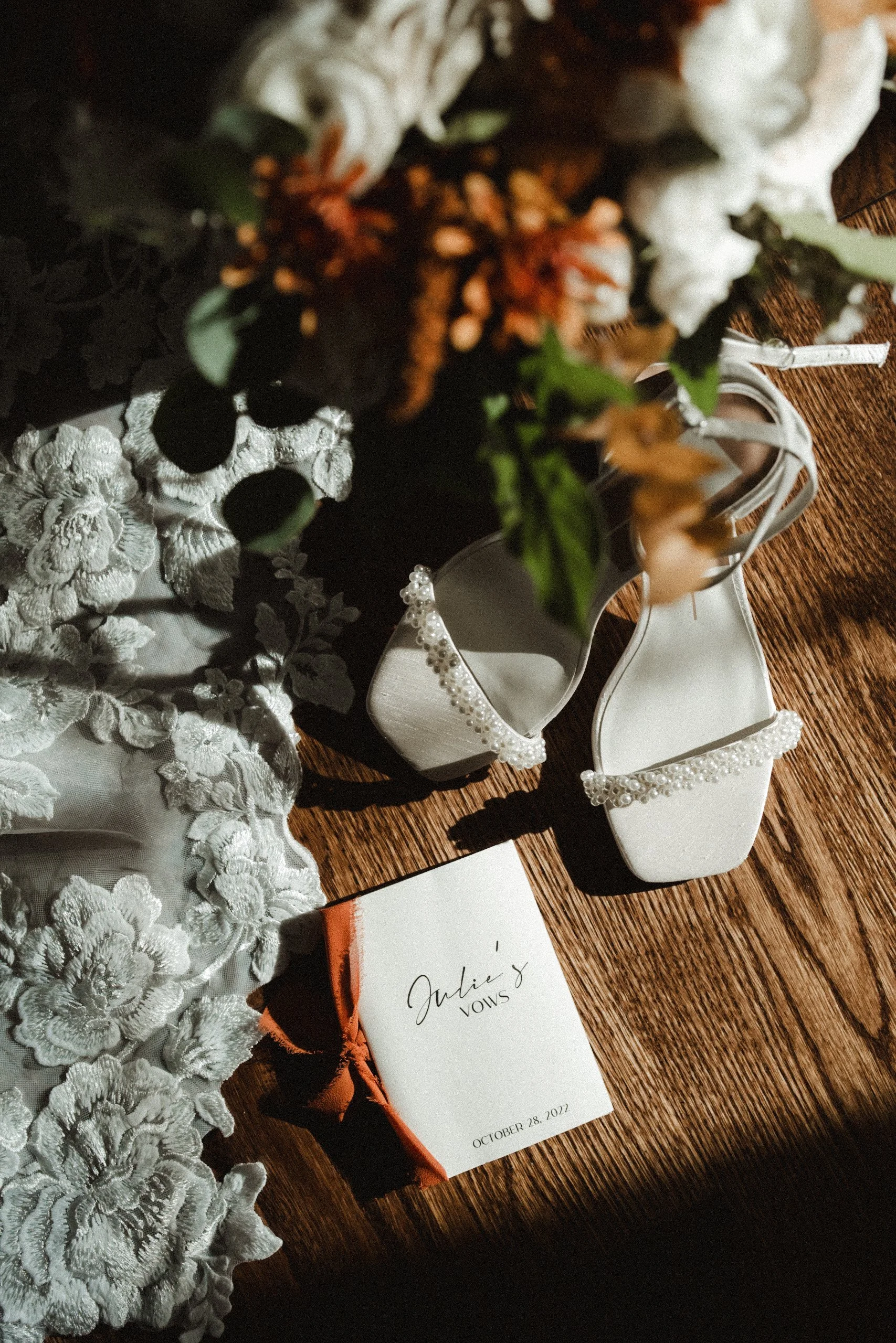 Wedding shoes with pearl embellishments, a bridal vow card with the name 'Julie's Vows' and date October 28, 2022, a floral bouquet, and lace fabric on a wooden surface.