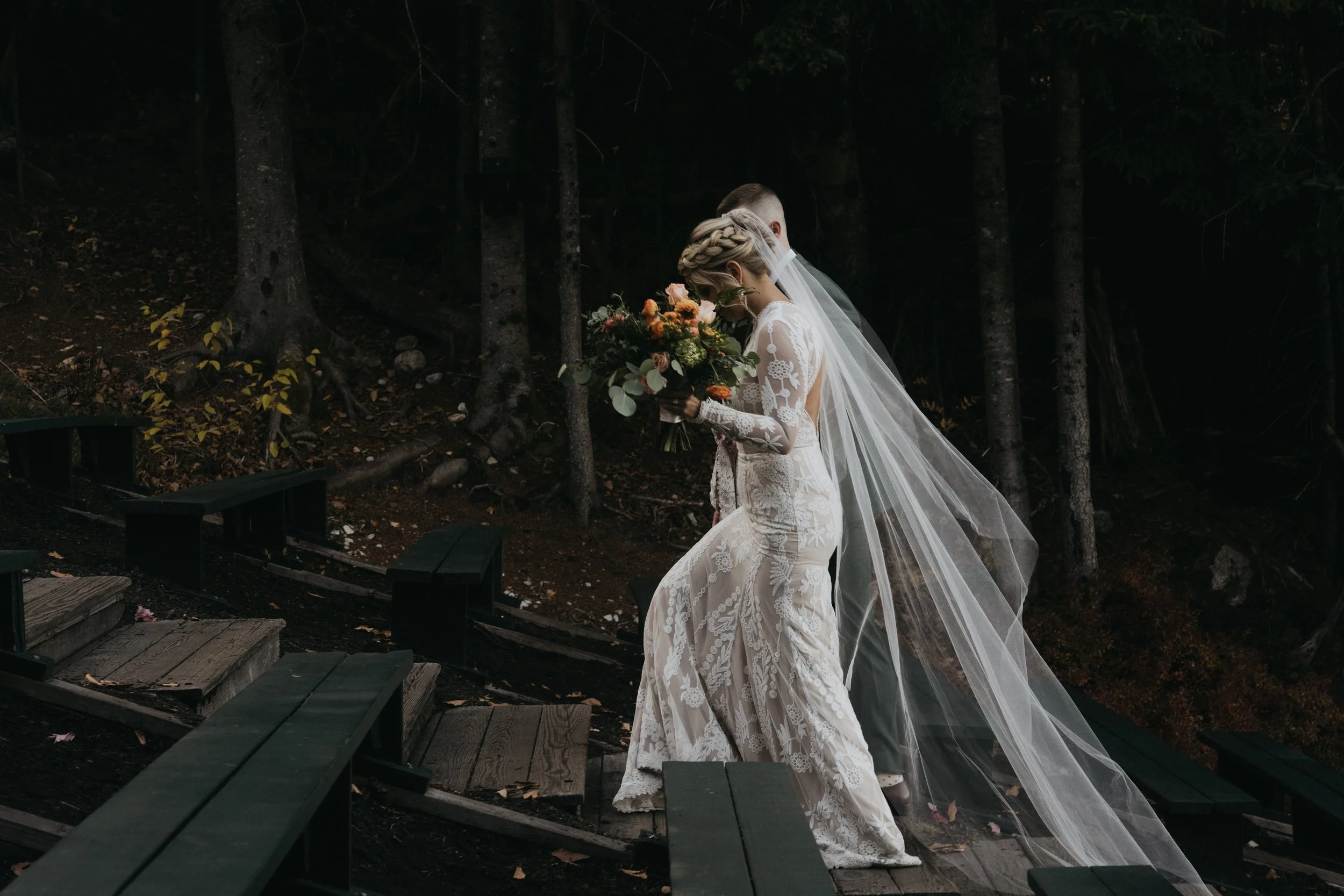 Bride in a lace wedding dress and veil holding a bouquet of flowers on a wooded outdoor staircase.
