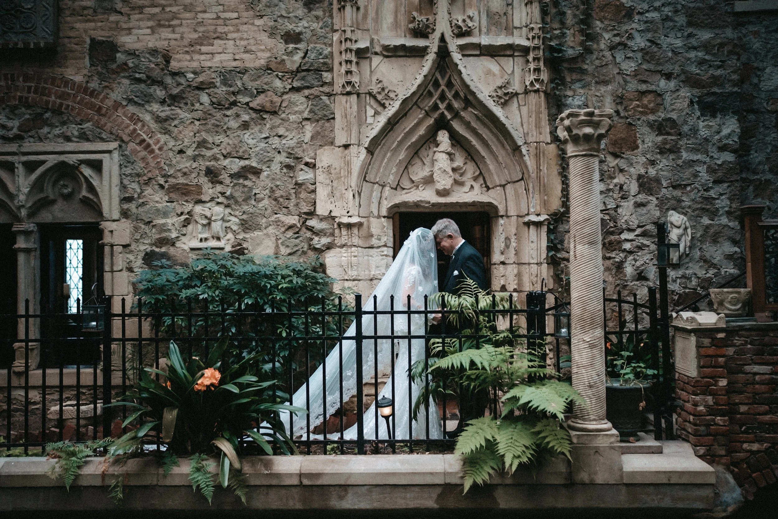 A bride and groom standing close together, holding hands, inside a stone chapel with gothic architecture, green plants, and decorative statues.