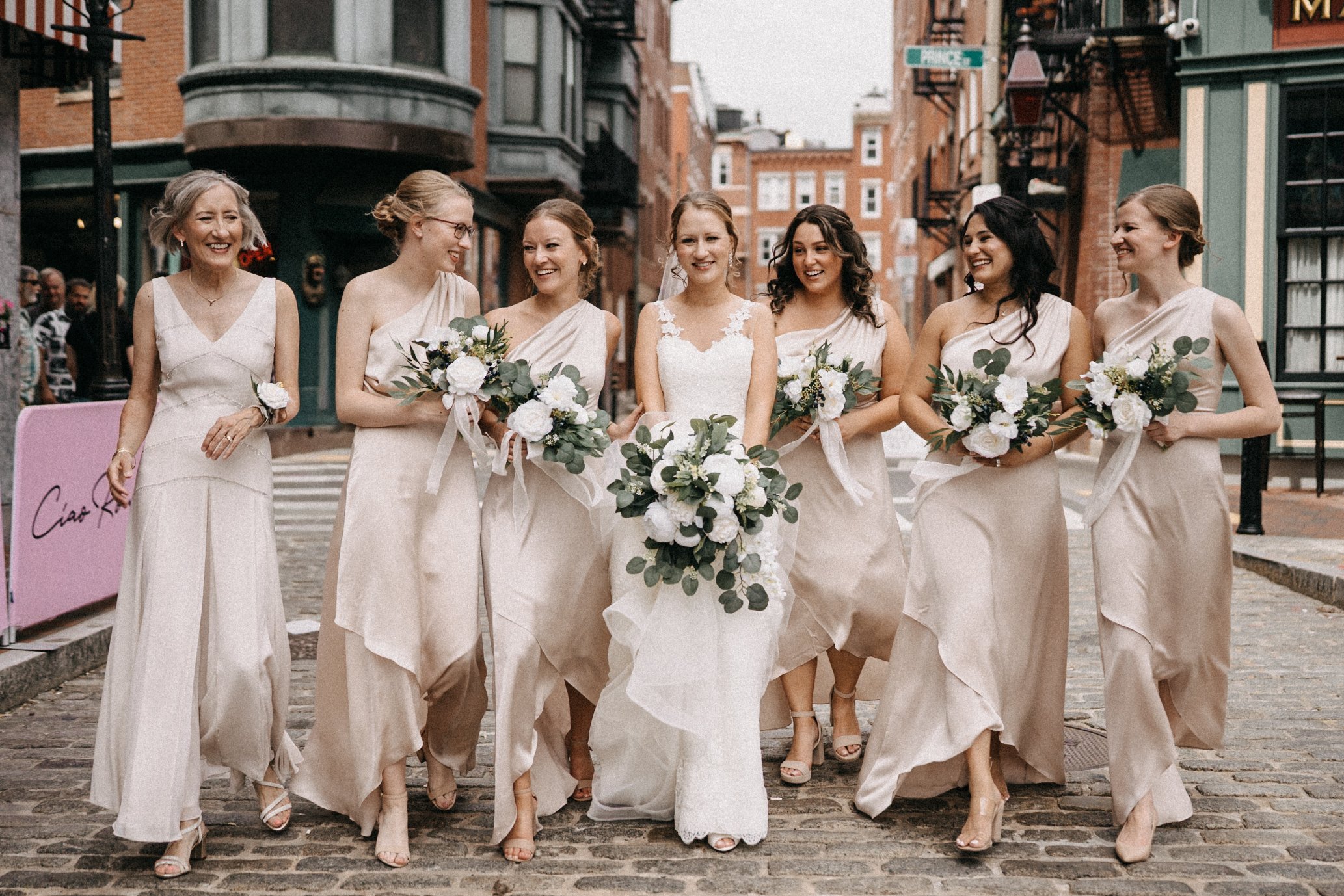 A group of seven women in beige dresses walking on a cobblestone street, smiling, holding bouquets, with buildings and a crosswalk in the background, celebrating a wedding.