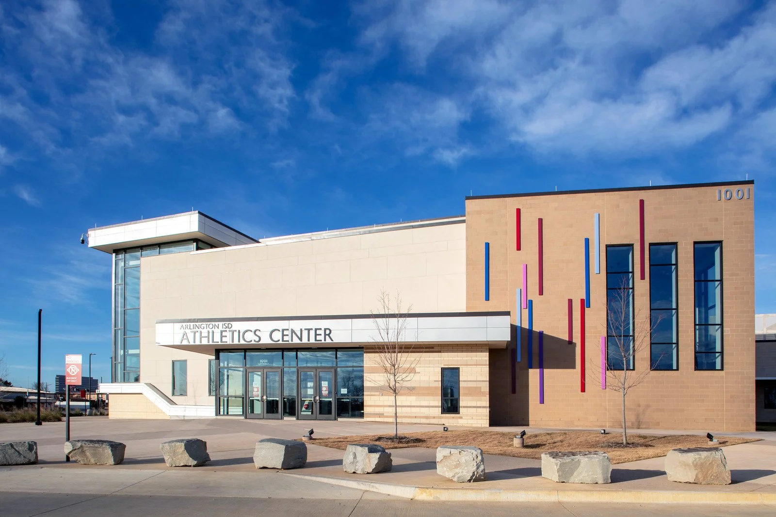 Exterior view of Arlington ISD Athletics Center, a modern building with beige and brown brick walls, colorful vertical art installation, large windows, and a clear blue sky.