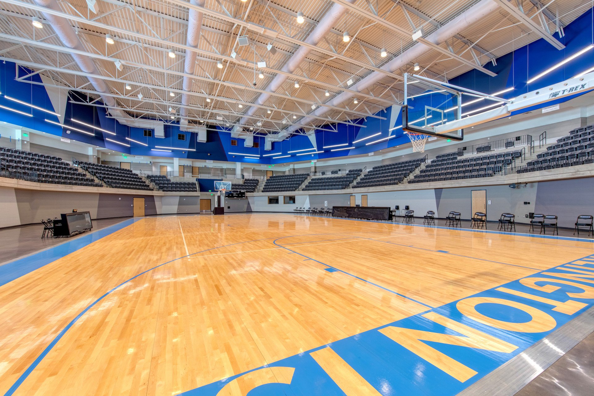 Empty indoor basketball court with polished wooden floor, blue and gray walls, and tiered seating around the court.