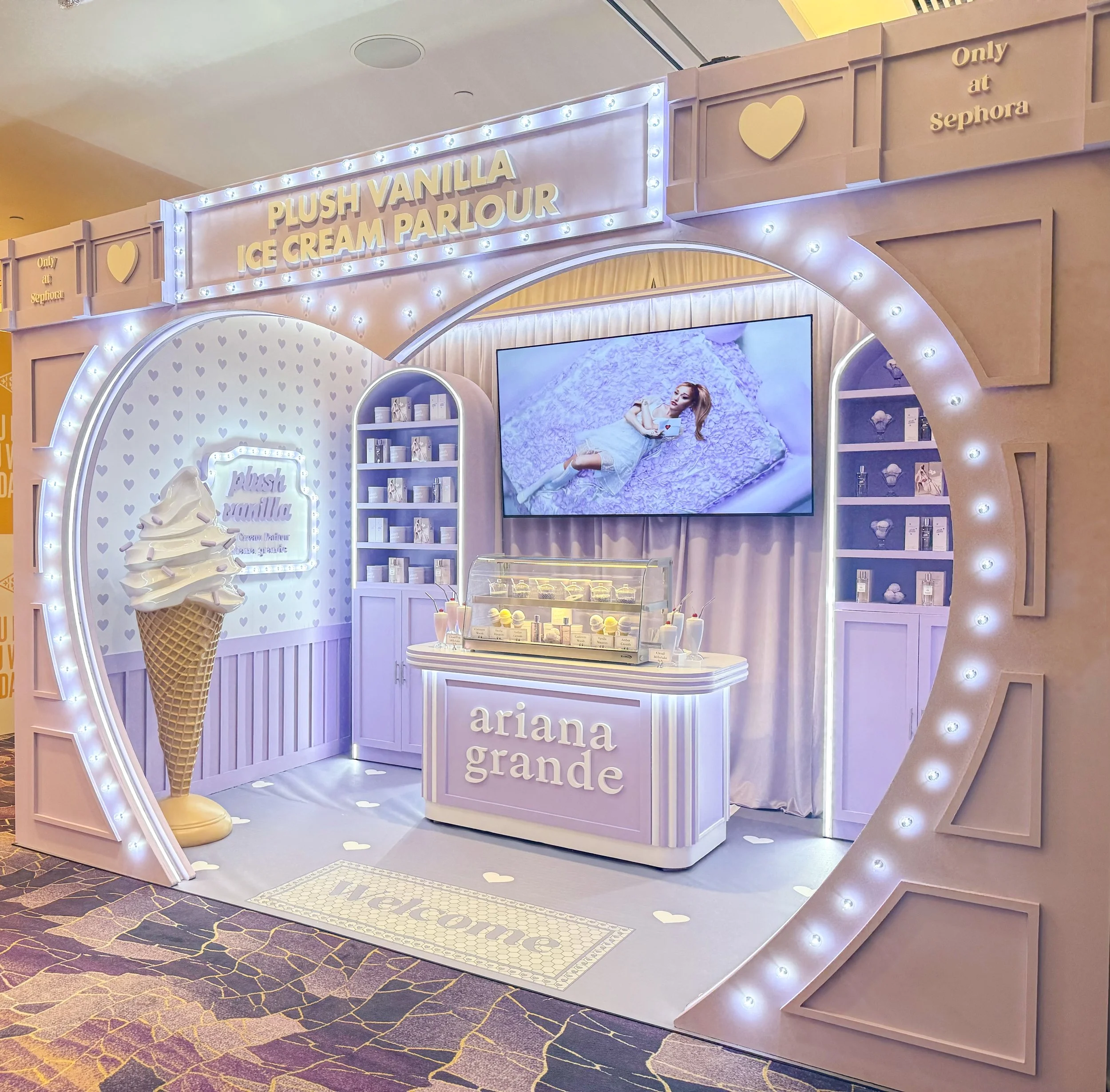 A decorated ice cream shop brand activation for the fragrance Plush Vanilla with pastel purple colors, a large ice cream cone, a TV showing Ariana Grande in a photoshoot, and signage that says 'Plush Vanilla Ice Cream Parlour' and 'Ariana Grande.'