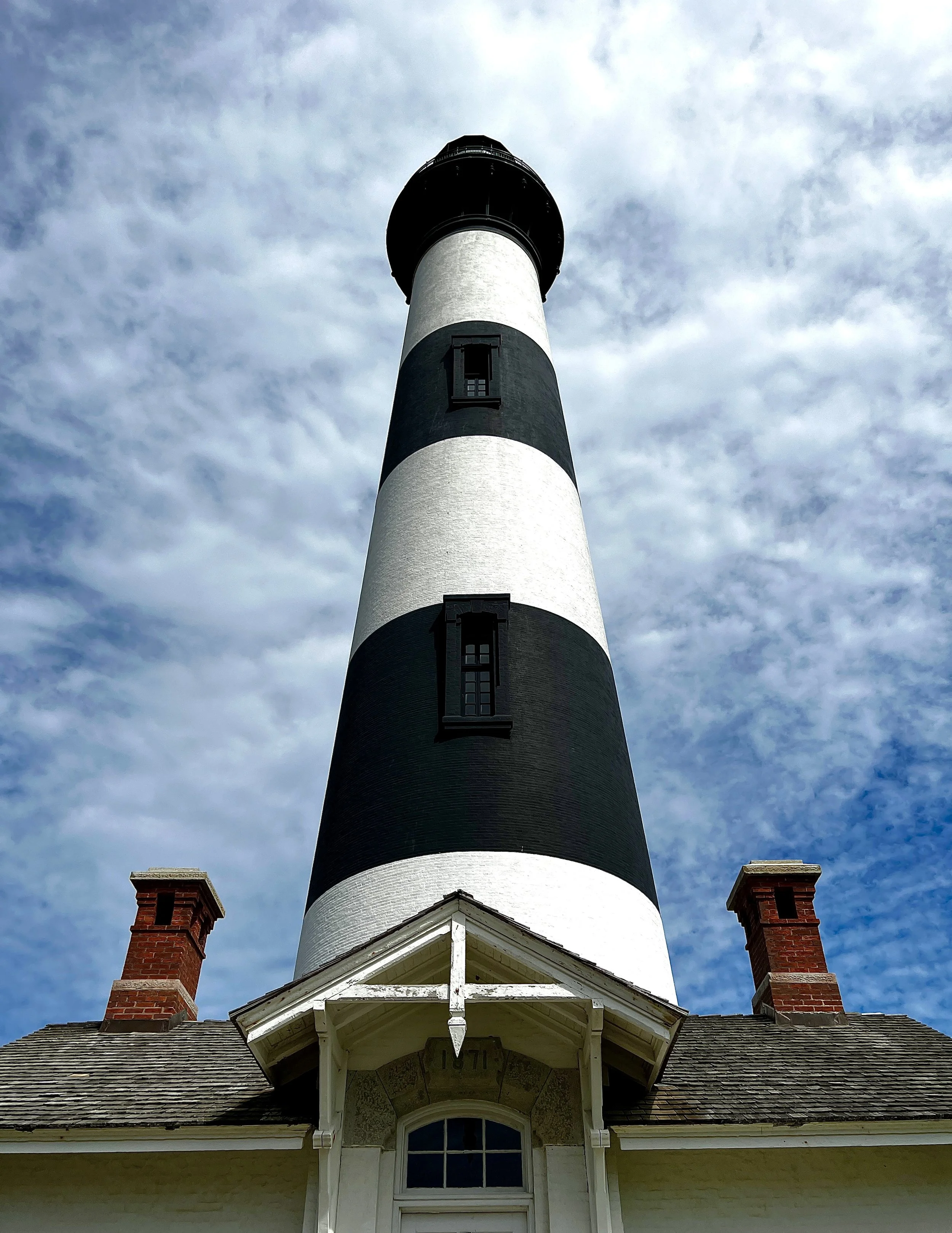 A tall, black and white striped lighthouse viewed from below against a cloudy sky, with a building at its base featuring a peaked roof and brick chimneys.