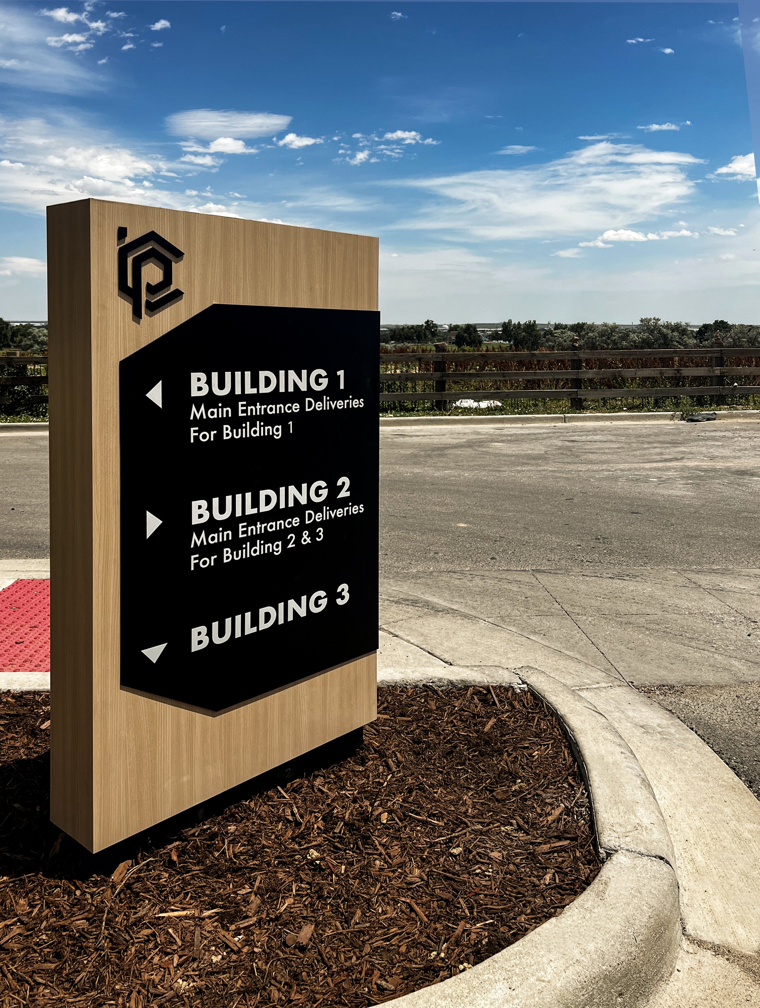 Wayfinding directional signpost with directions to three buildings and their delivery areas in an outdoor parking lot, with a blue sky and distant trees in the background.