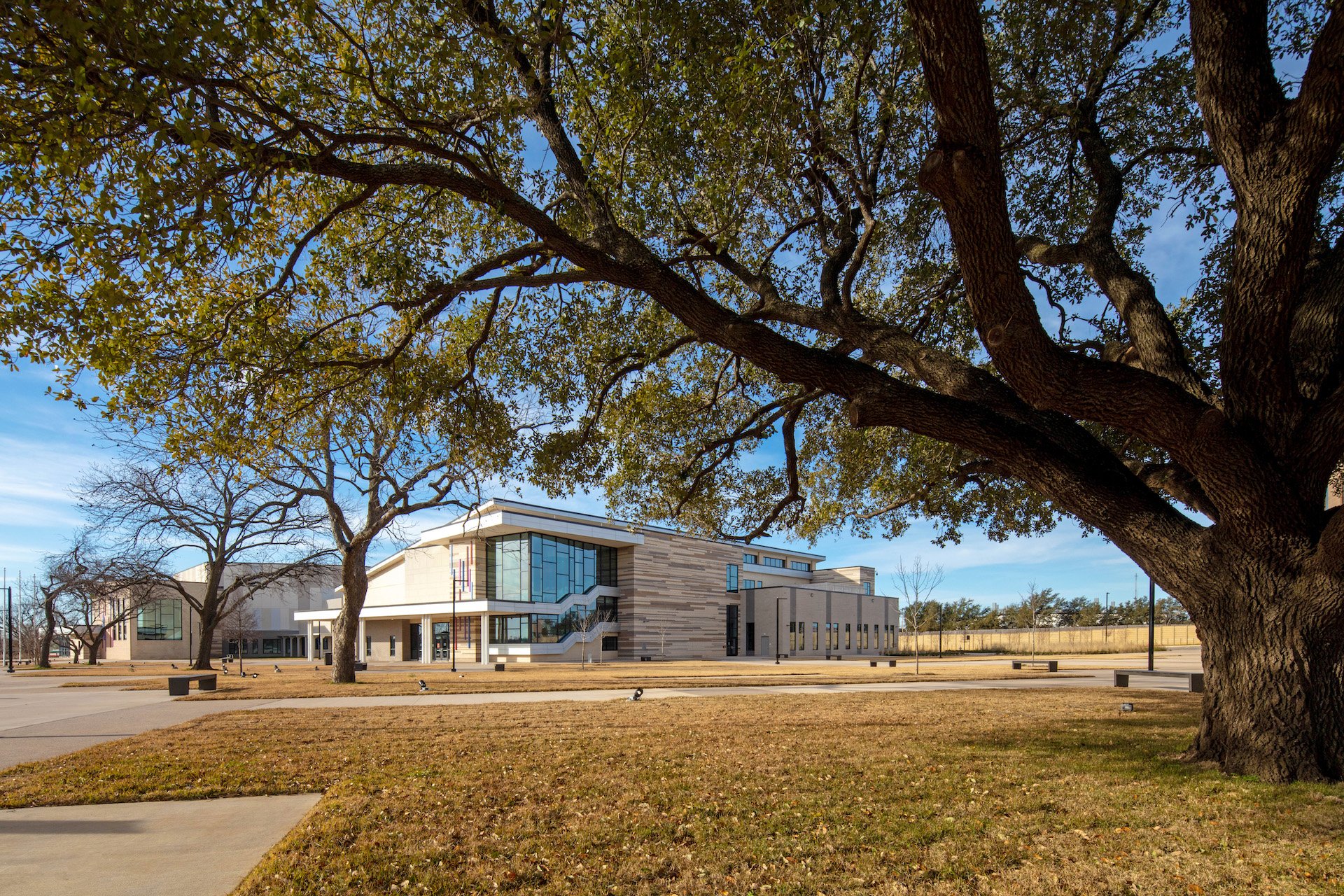 A modern building with large glass windows and a beige exterior, surrounded by leafless trees, under a partly cloudy sky.