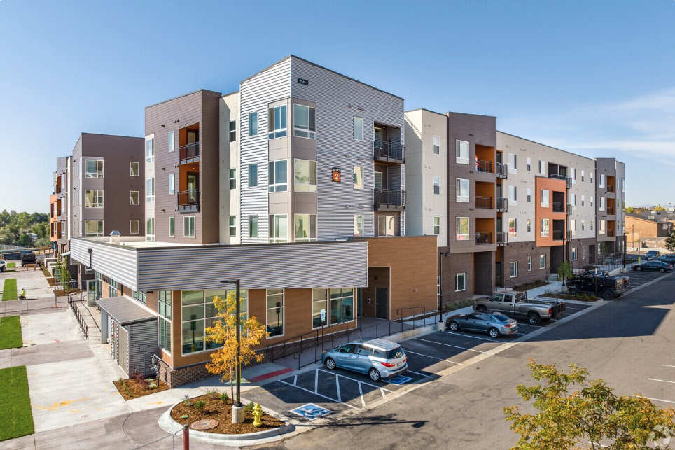 Modern multi-story apartment building with parking lot in front, clear blue sky, and surrounding trees.