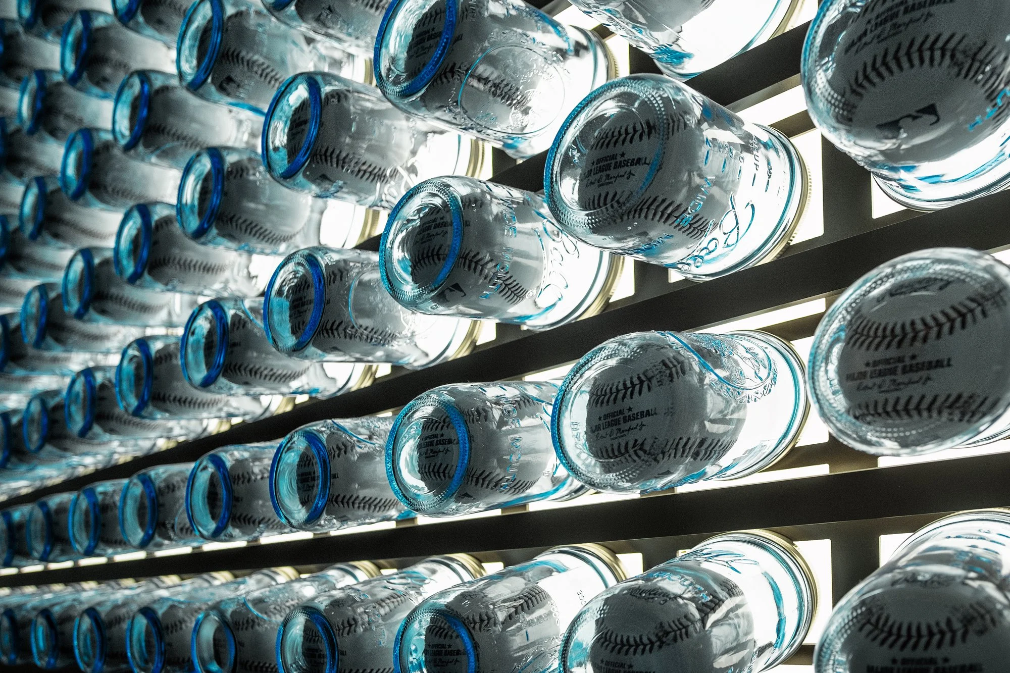 Multiple transparent glass mason jars with blue caps, stacked on metal shelves against a backlit LED display for the MLB Milwaukee Brewers locker room art moments. 
