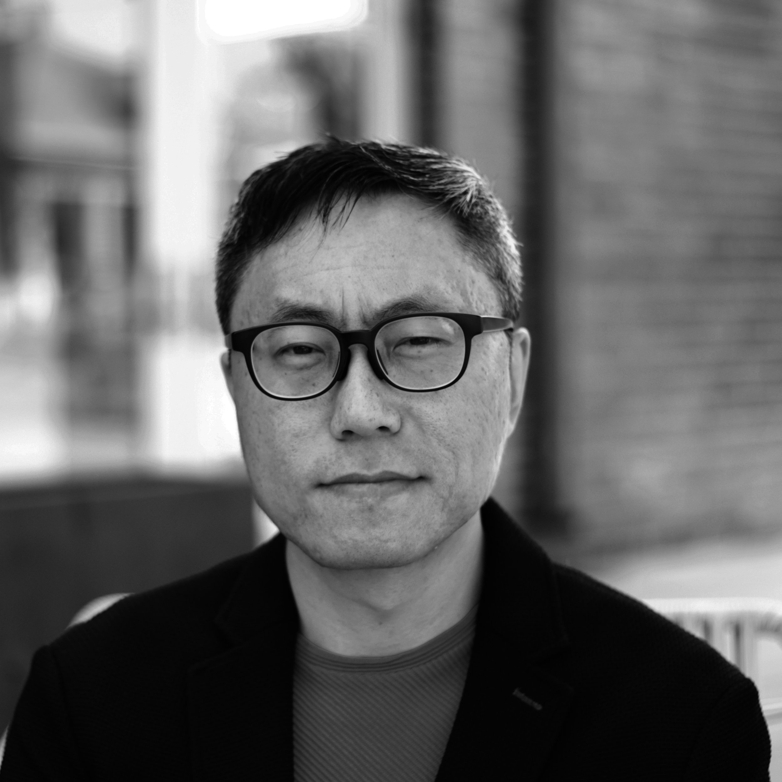 A man with glasses and short dark hair, wearing a blazer and shirt, looking at the camera in an indoor setting.