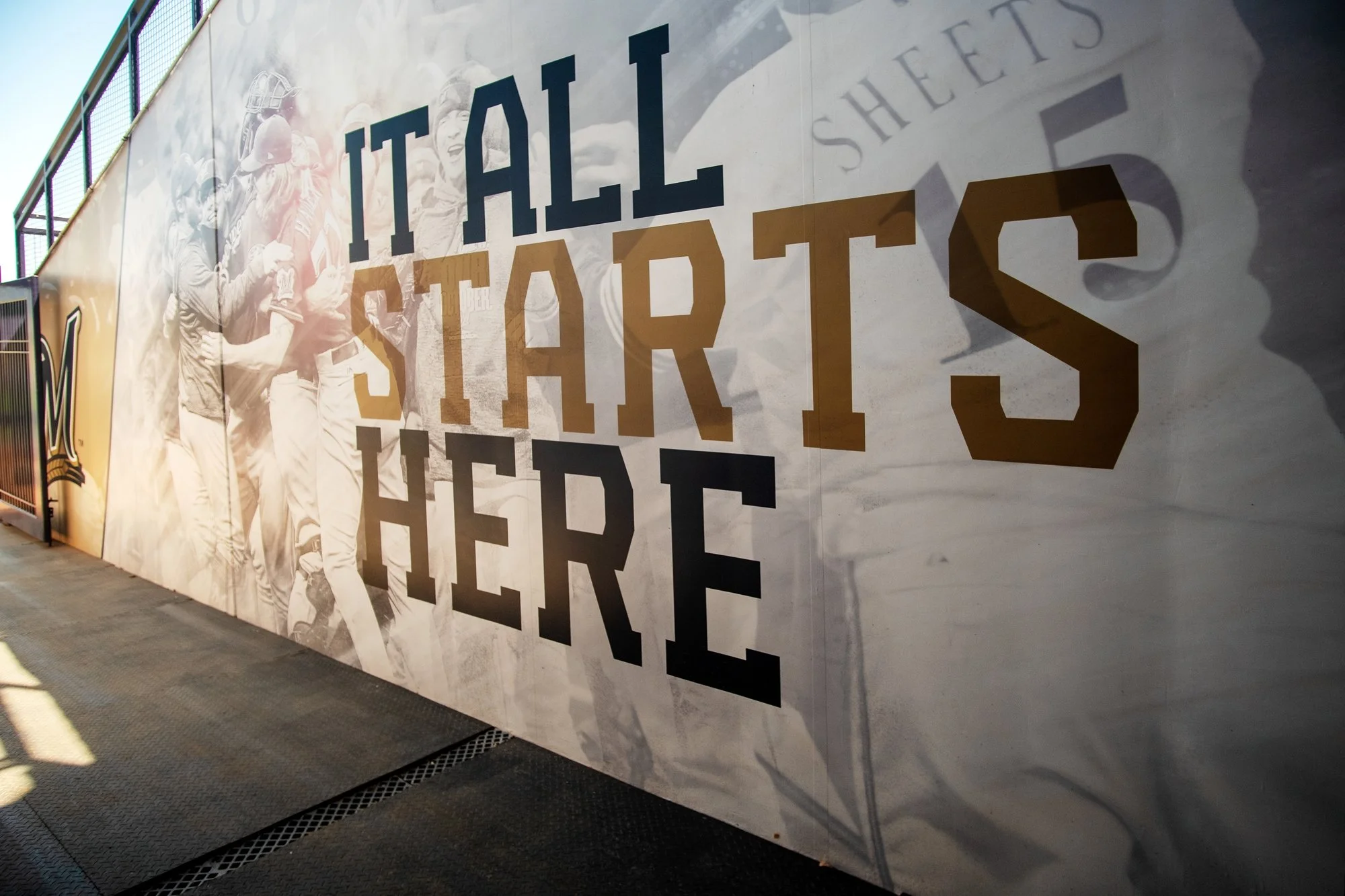 A large scale wall graphic for the hype tunnel with a grayscale image of MLB Milwaukee Brewers baseball players and the words 'IT ALL STARTS HERE' in big black and tan letters.