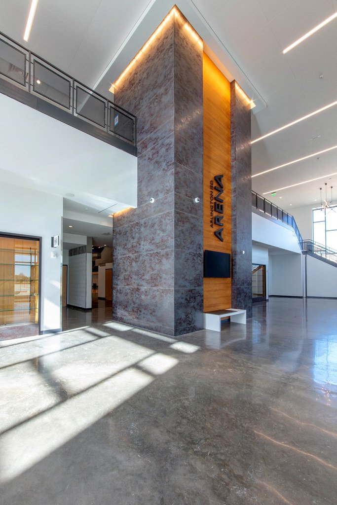 Interior of a modern office building lobby with a large central stone column featuring a wooden panel with the company name "AIREX" and a digital screen, bright lighting, polished concrete floors, and a glass balcony on the upper level.
