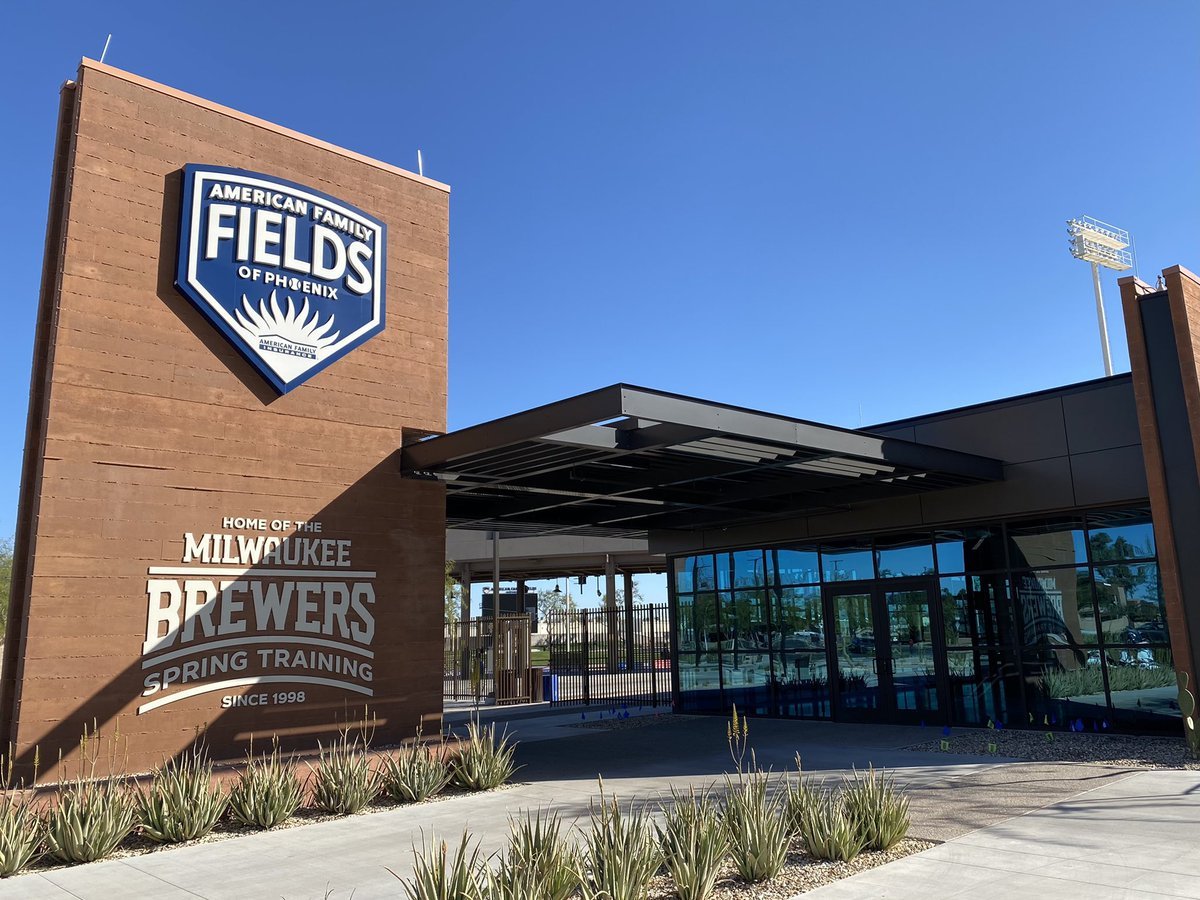 The entrance of American Family Fields of Phoenix baseball stadium with signage indicating it is the home of the Milwaukee Brewers spring training, featuring a brick wall with a large blue and white sign, and a glass door entrance, under a clear blue