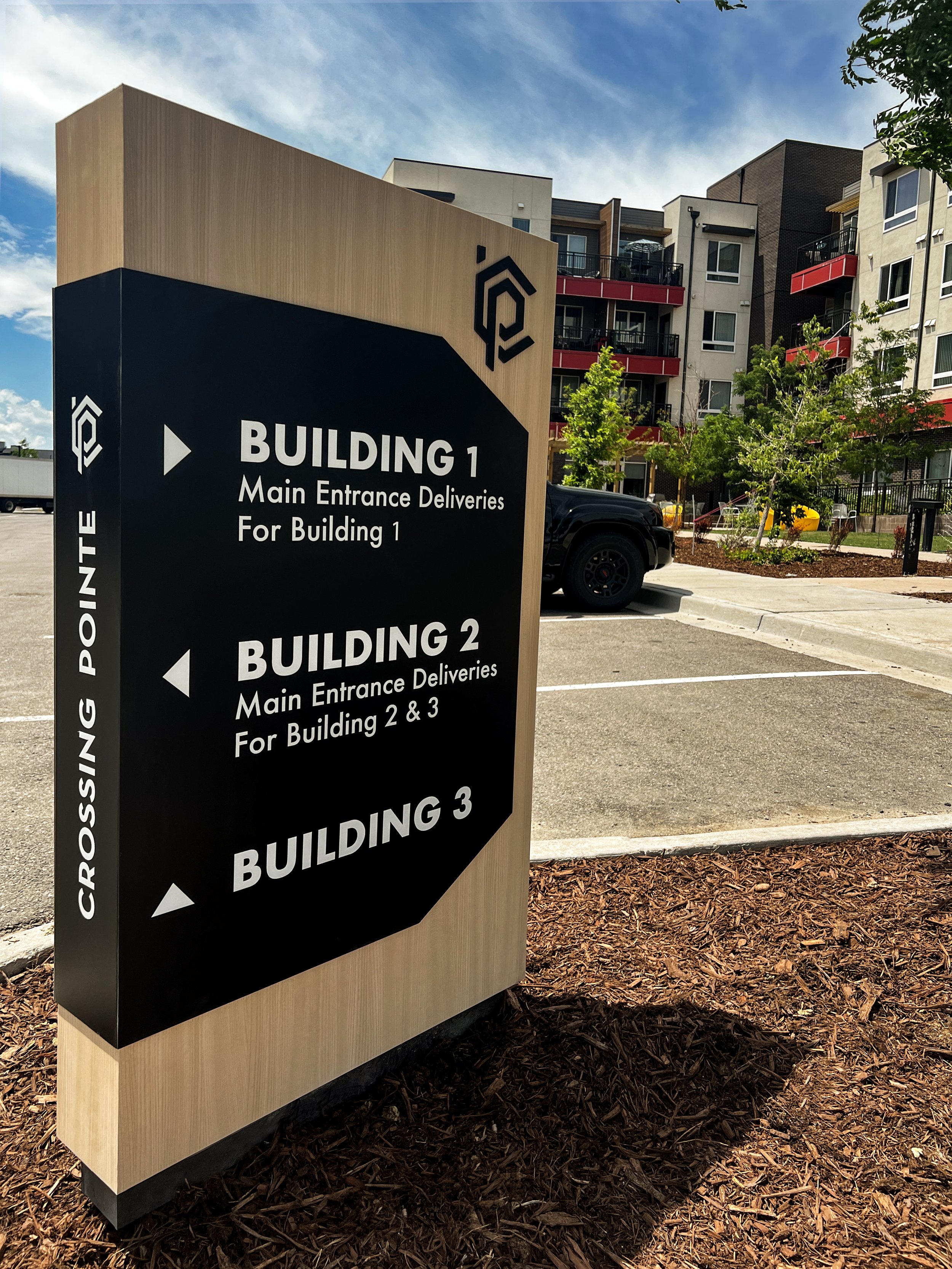 Sign indicating directions to Building 1, Building 2, and Building 3, with main entrance deliveries for each building, situated outdoors in a parking lot with residential apartment buildings in the background.