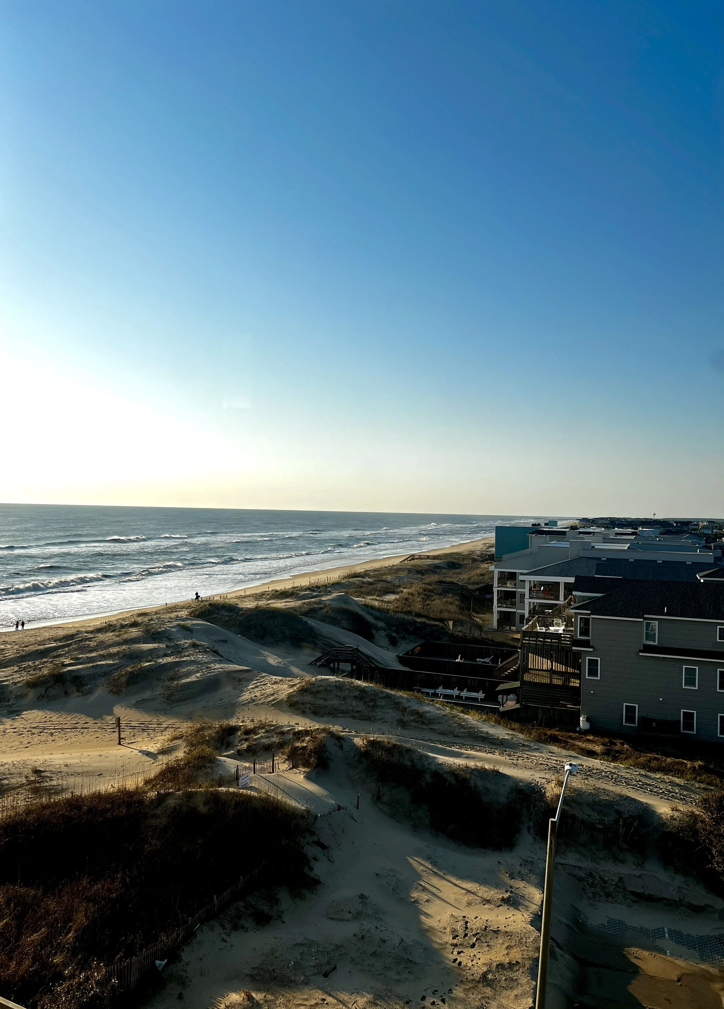 View of a beach with sand dunes, ocean waves, and residential buildings along the coast under a clear blue sky during sunset or sunrise.