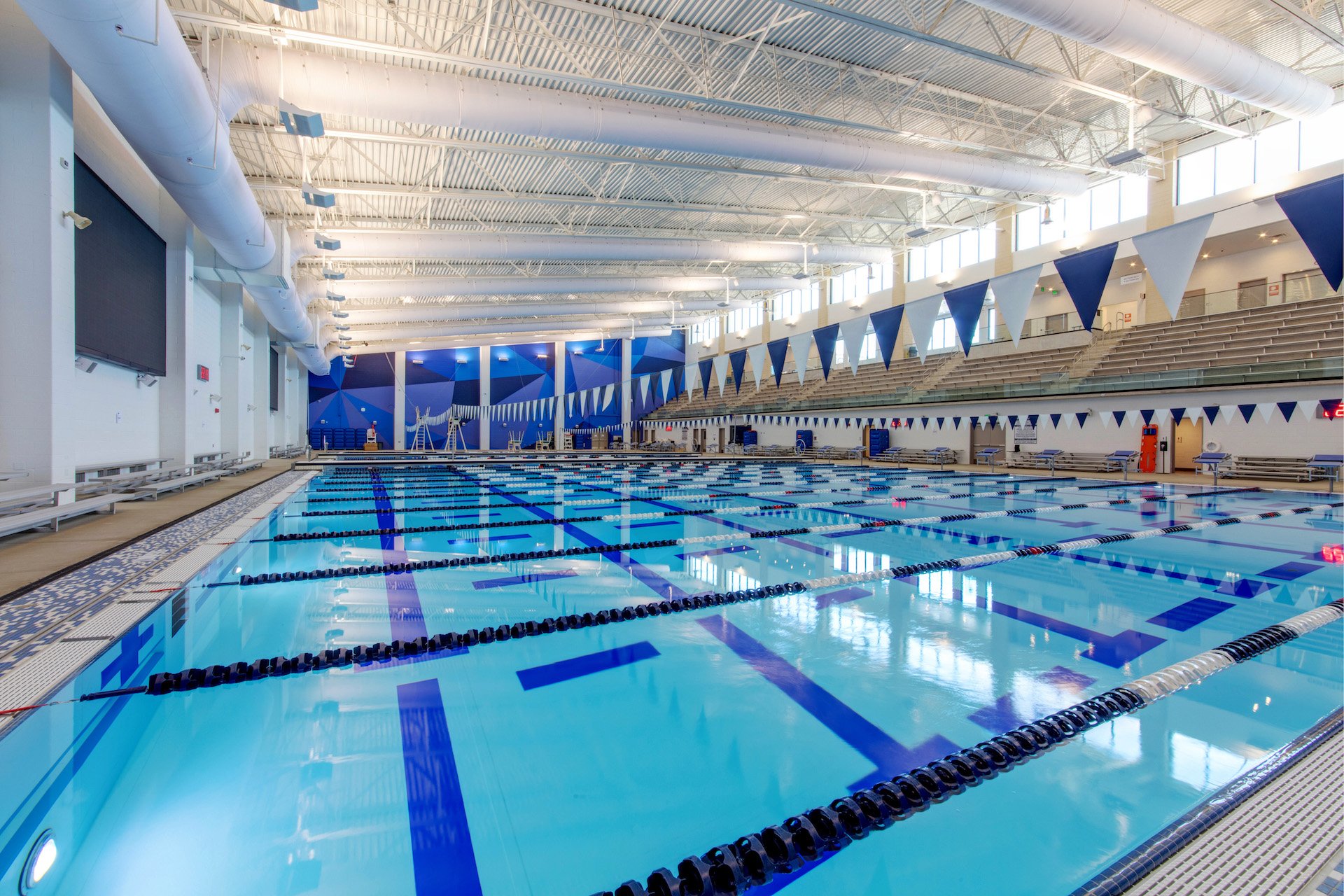 Indoor swimming pool with multiple lanes, blue and white flags hanging overhead, and large windows allowing natural light. Large vinyl graphics are on the opposite wall for photo ops. 