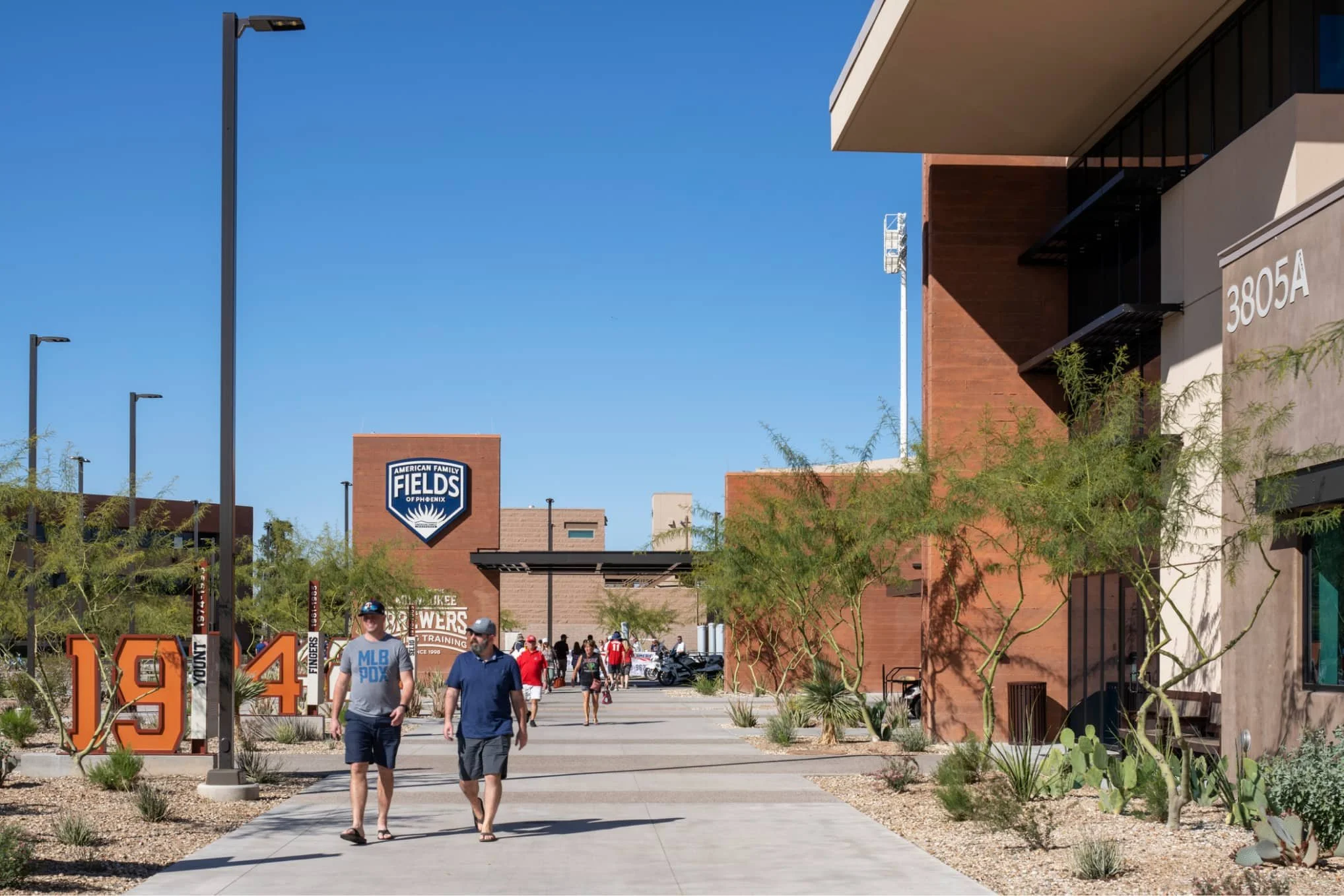 People walking on a pathway in a sports complex with modern buildings, trees, and desert plants. A sign reads 'American Family Fields of Phoenix' in the background of the MLB Milwaukee Brewers spring training facility.