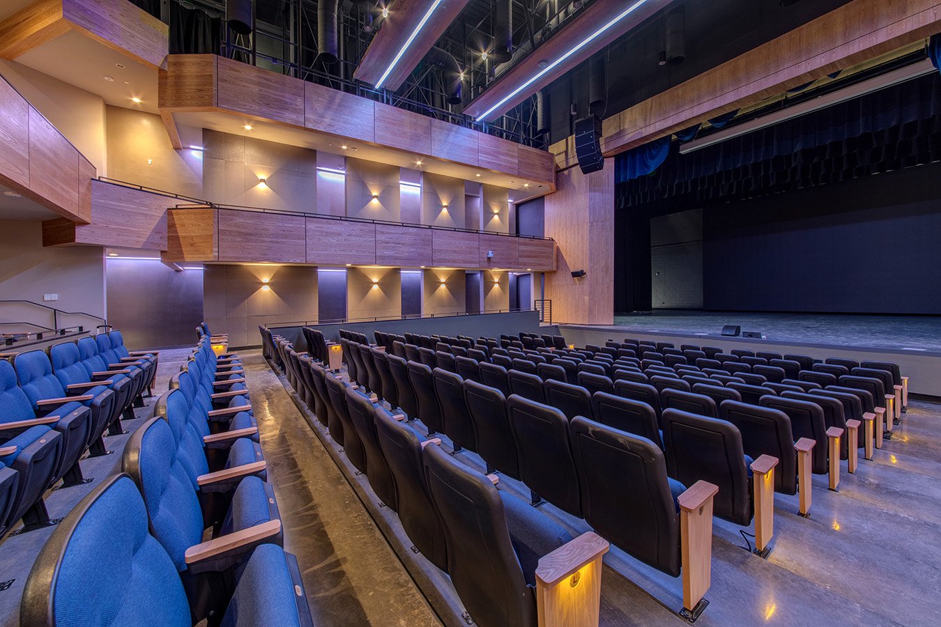 Empty theater with blue and black seats, wooden accents, and a large stage with curtains.
