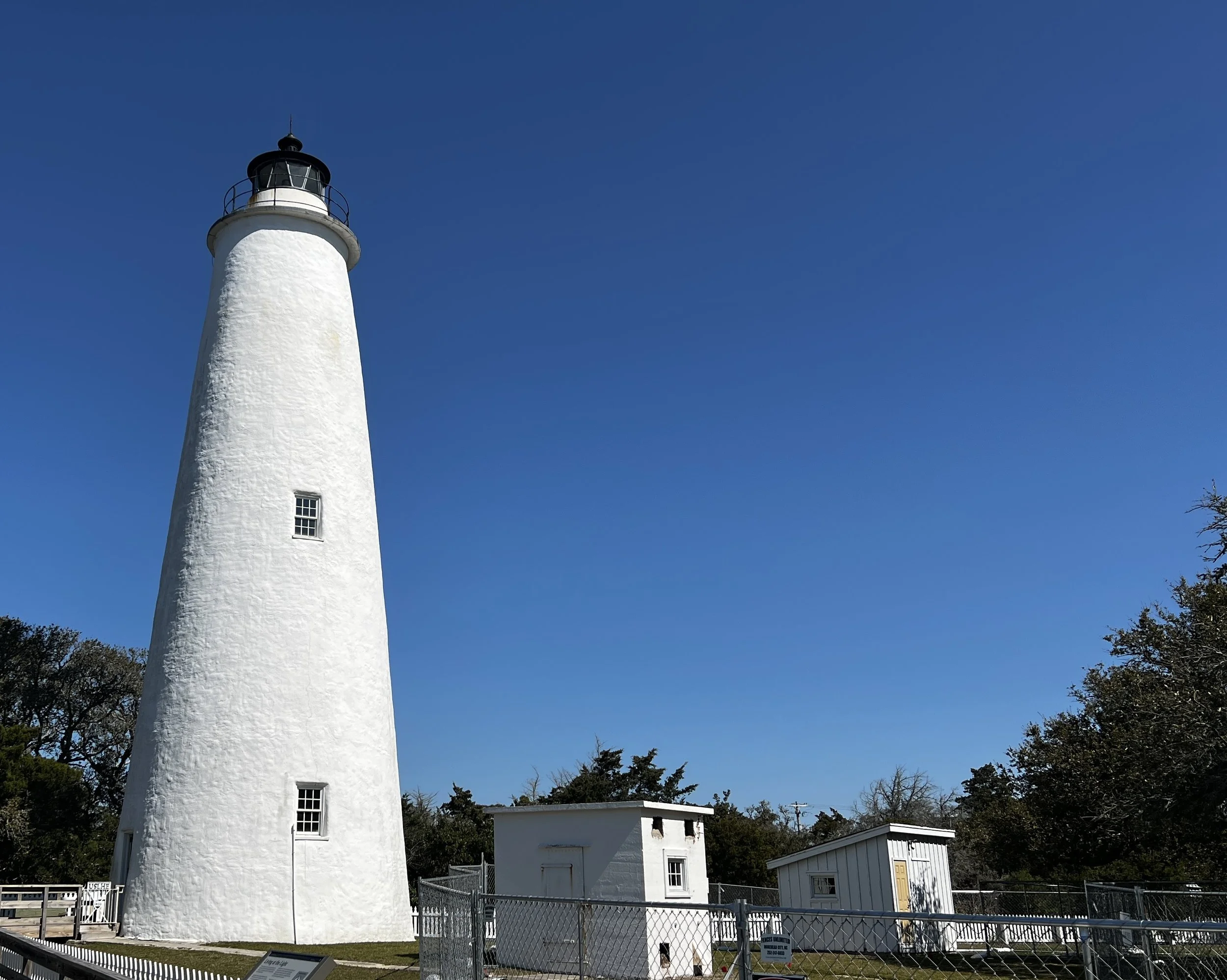 A white lighthouse with a black top under a clear blue sky, surrounded by small white buildings and fenced area.