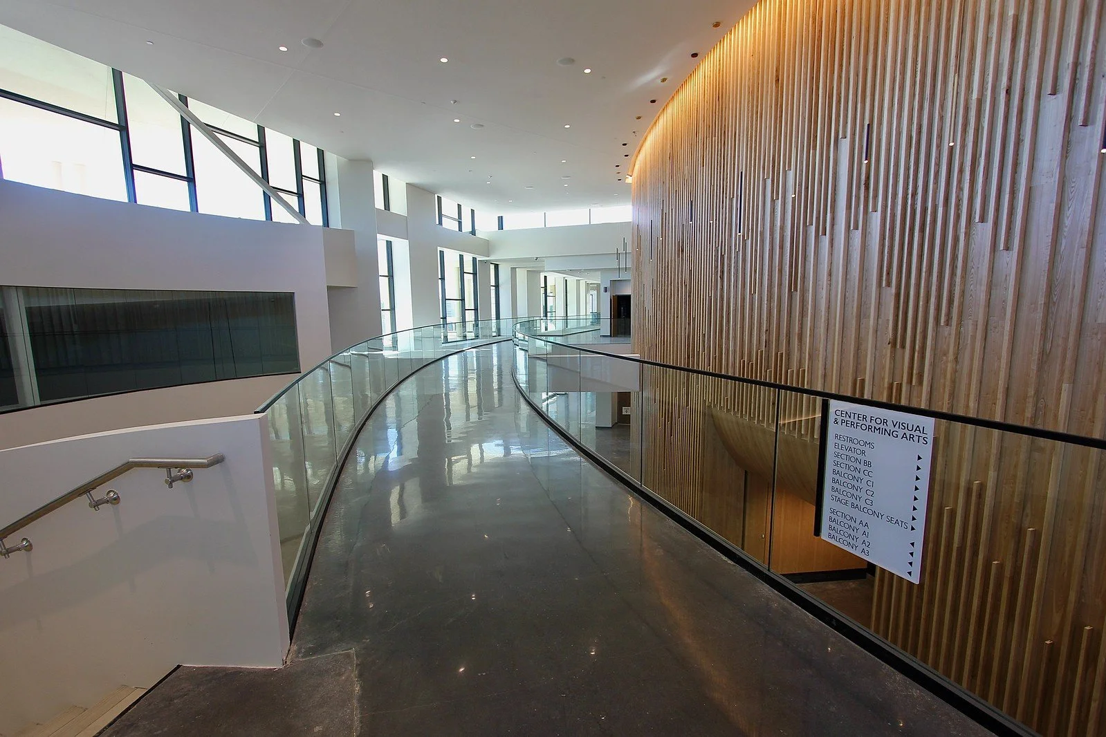 Interior view of a modern building with a curved glass railing, large windows, and a wooden wall with a sign listing various sections and amenities.