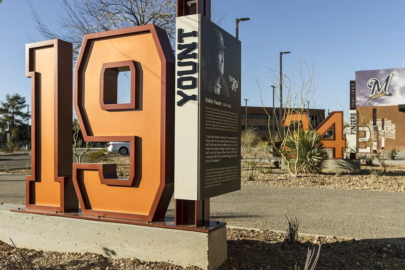 Large orange retired player jersey number '94' with an informational plaque in front, set in a desert landscape with leafless trees and buildings in the background for the MLB Milwaukee Brewers facility. 