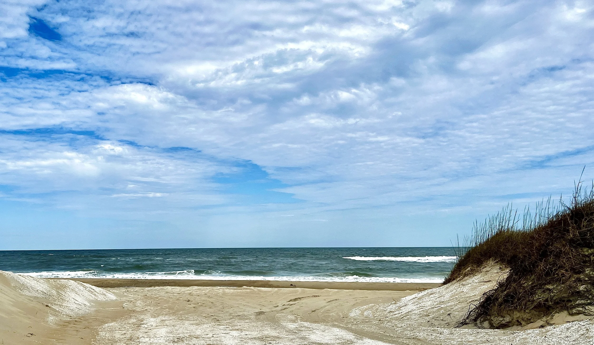 A sandy beach with tire tracks, grassy dunes on the right, and an ocean with small waves under a cloudy sky.
