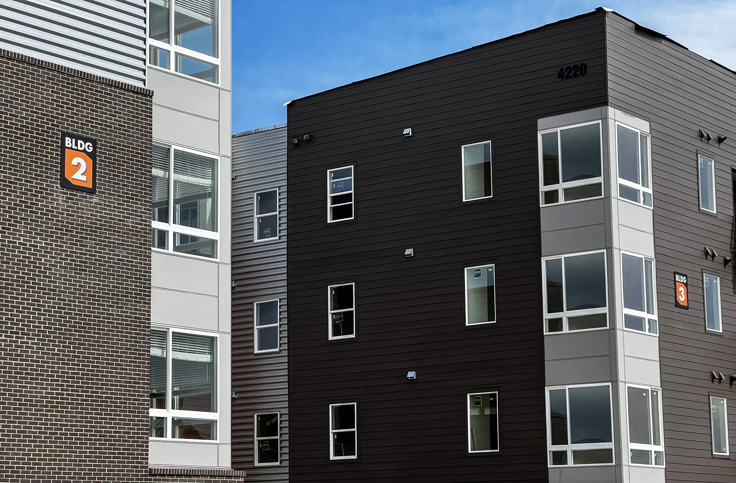 Multiple modern apartment buildings with brick, metal, and glass exteriors, blue sky, and building labels indicating BLDG 2 and BLDG 3.
