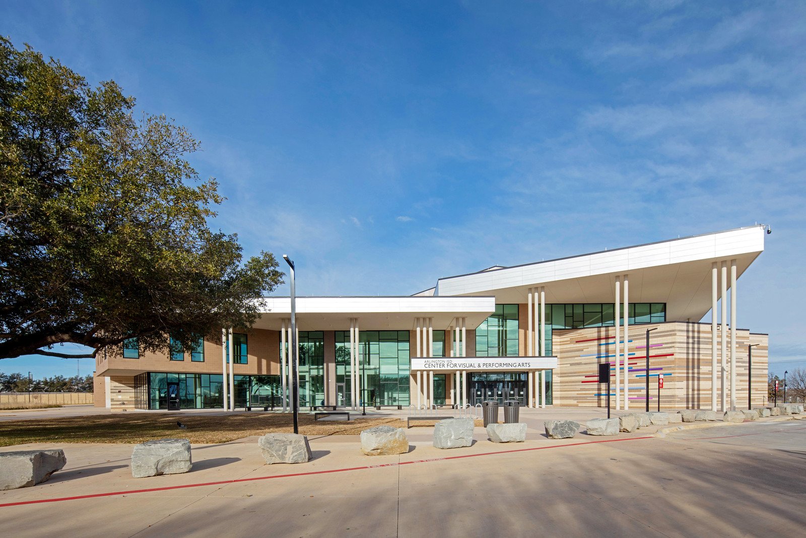 Modern building with large glass windows, white canopy, decorative colored stripes on the wall, and a sign indicating it is the Center for Visual & Performing Arts, with a clear blue sky and some large rocks in the foreground.