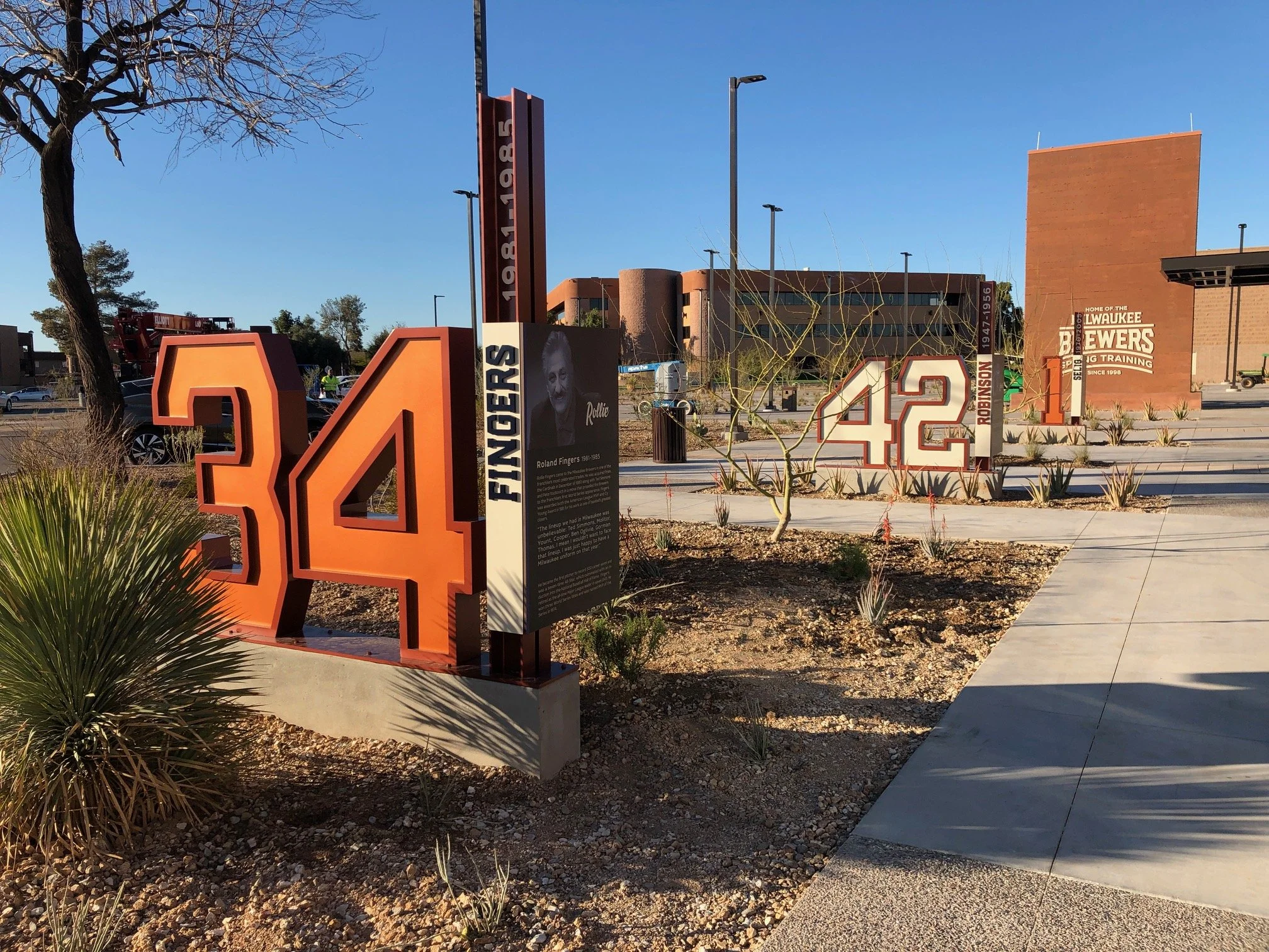 A series of large orange and white numbers displayed outdoors, with the number 34 in the foreground, followed by 42 and 1 in the background, on a sidewalk with small plants and desert landscaping.