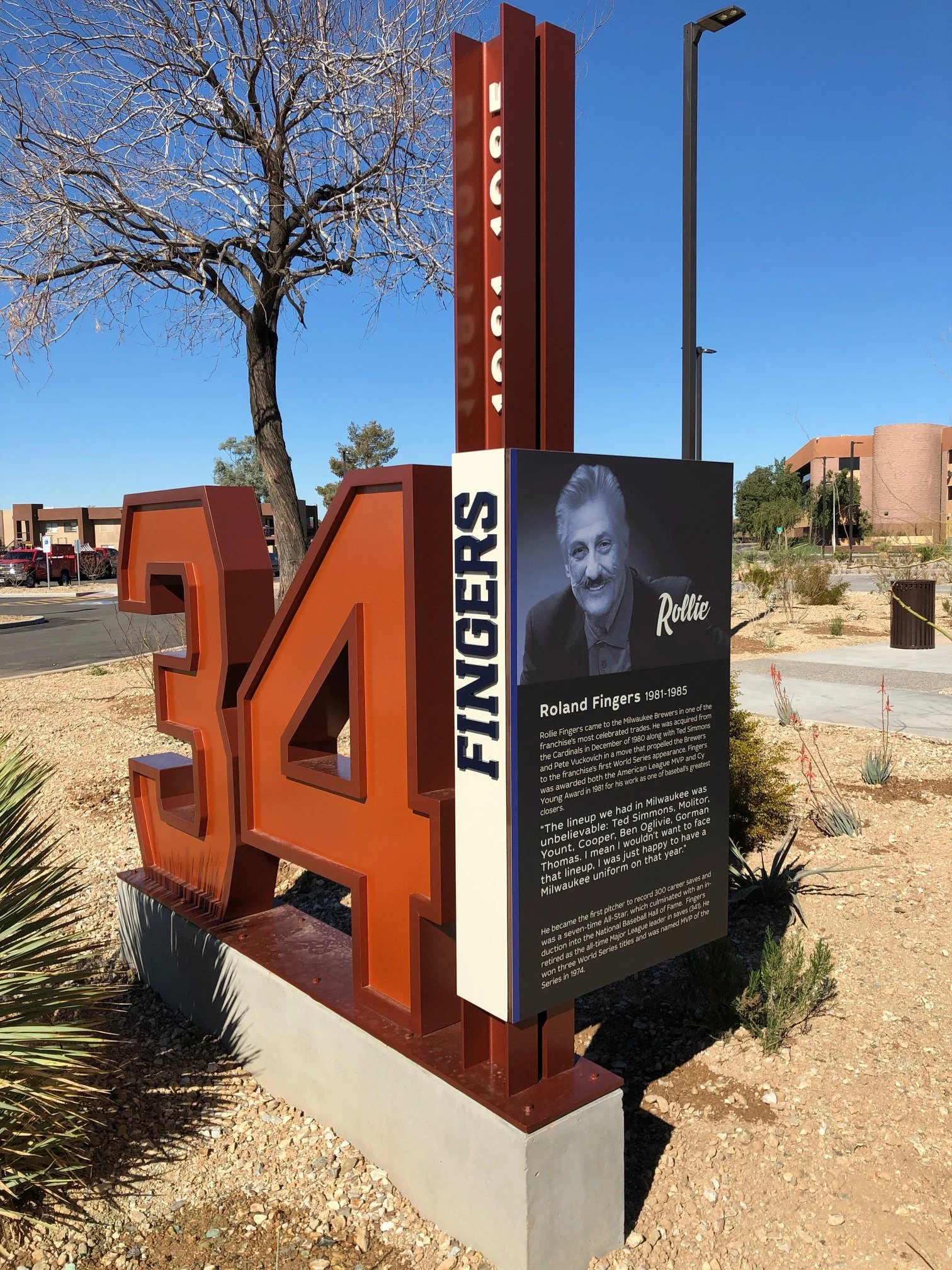A memorial display dedicated to Rollie Fingers featuring a large orange number 34, a black and white photo of Rollie Fingers, and a descriptive plaque about his career achievements in baseball, set outdoors on a sunny day with a clear blue sky, trees