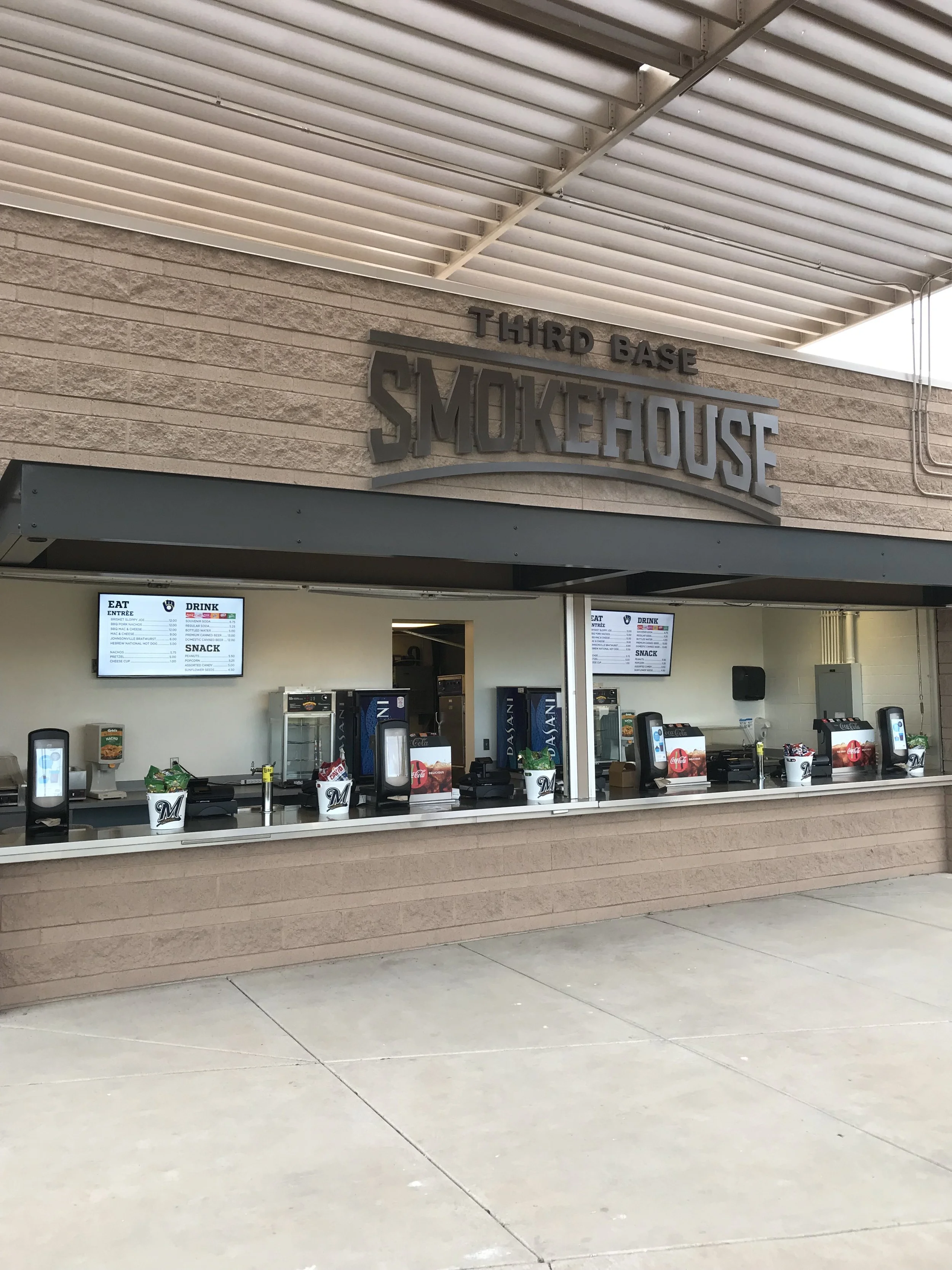 Outdoor counter at Third Base Smokehouse restaurant with digital menu boards, drink dispensers, and empty seats.