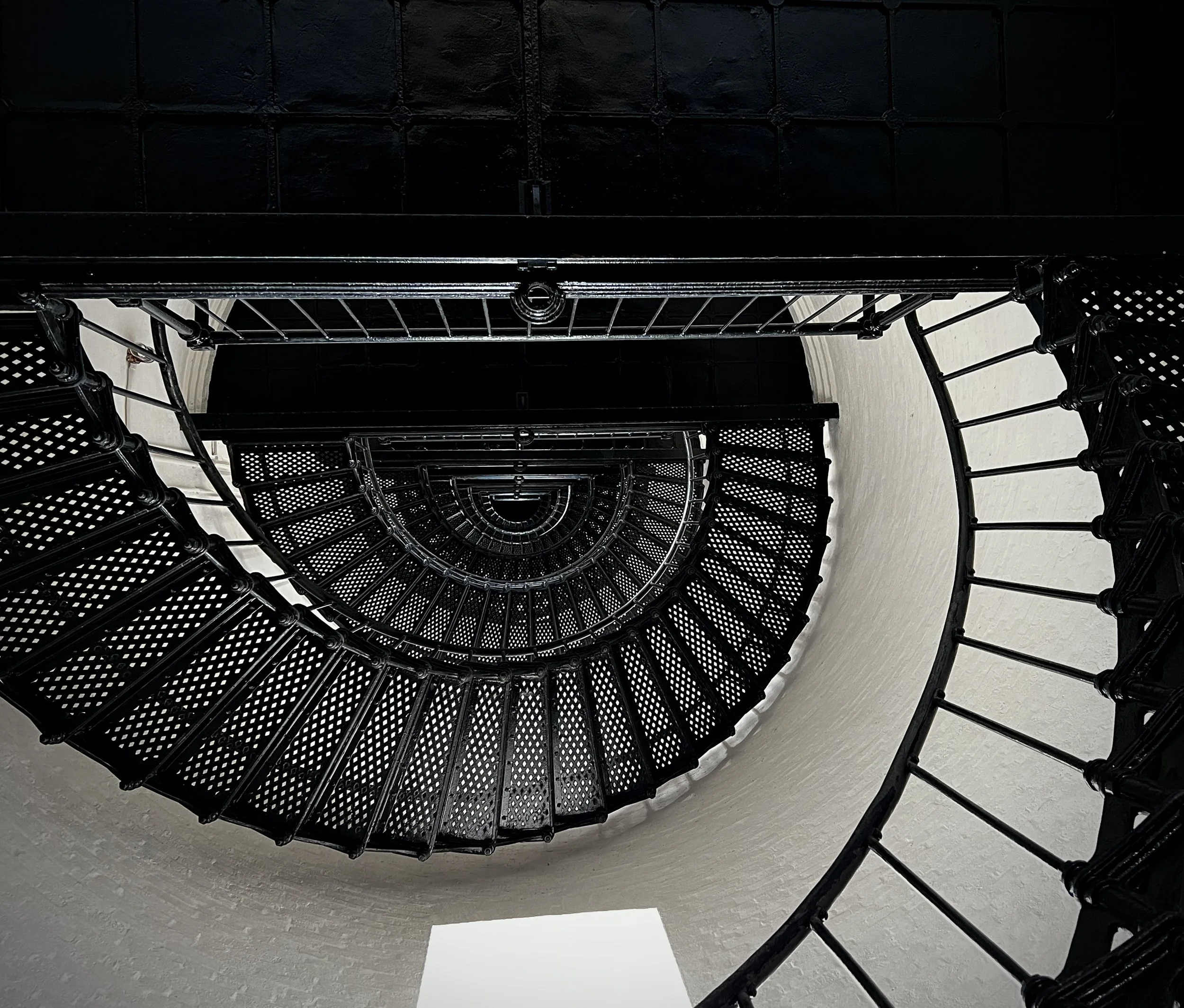 A top-down view of a spiral staircase with black metal railings and stairs, descending into a white cylindrical space.