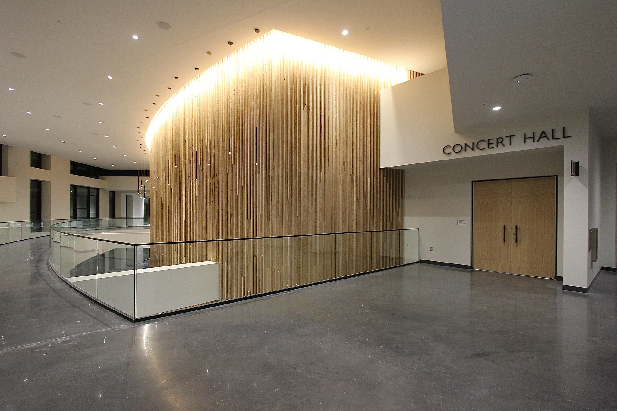 Interior of a modern concert hall with a curved wooden wall, glass railing, and a door labeled 'CONCERT HALL'.