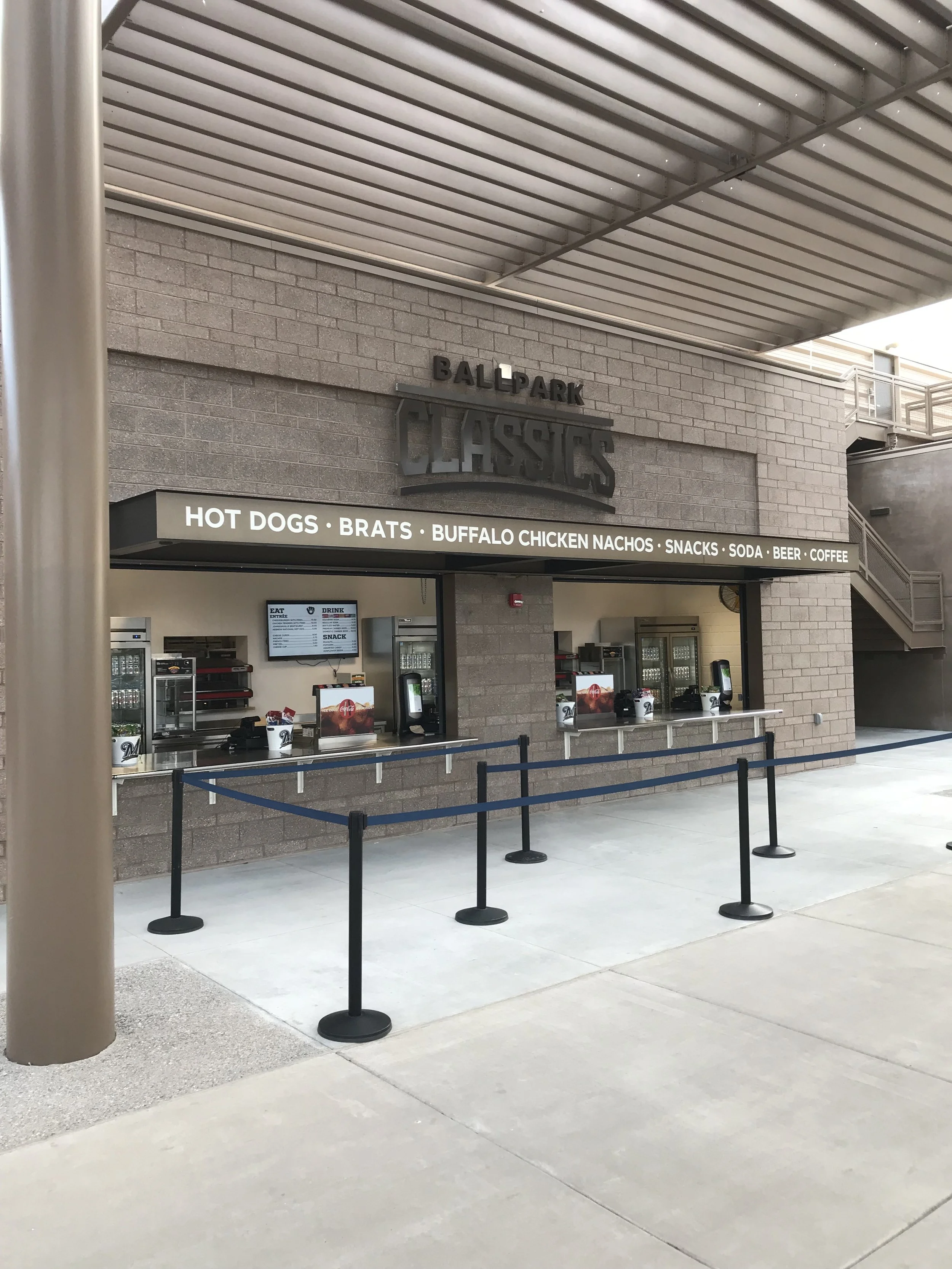 Outdoor concession stand with a sign reading 'Ballpark Classics' offering hot dogs, brats, buffalo chicken nachos, snacks, soda, beer, and coffee, with stanchions and a digital menu display for the MLB Milwaukee Brewers.