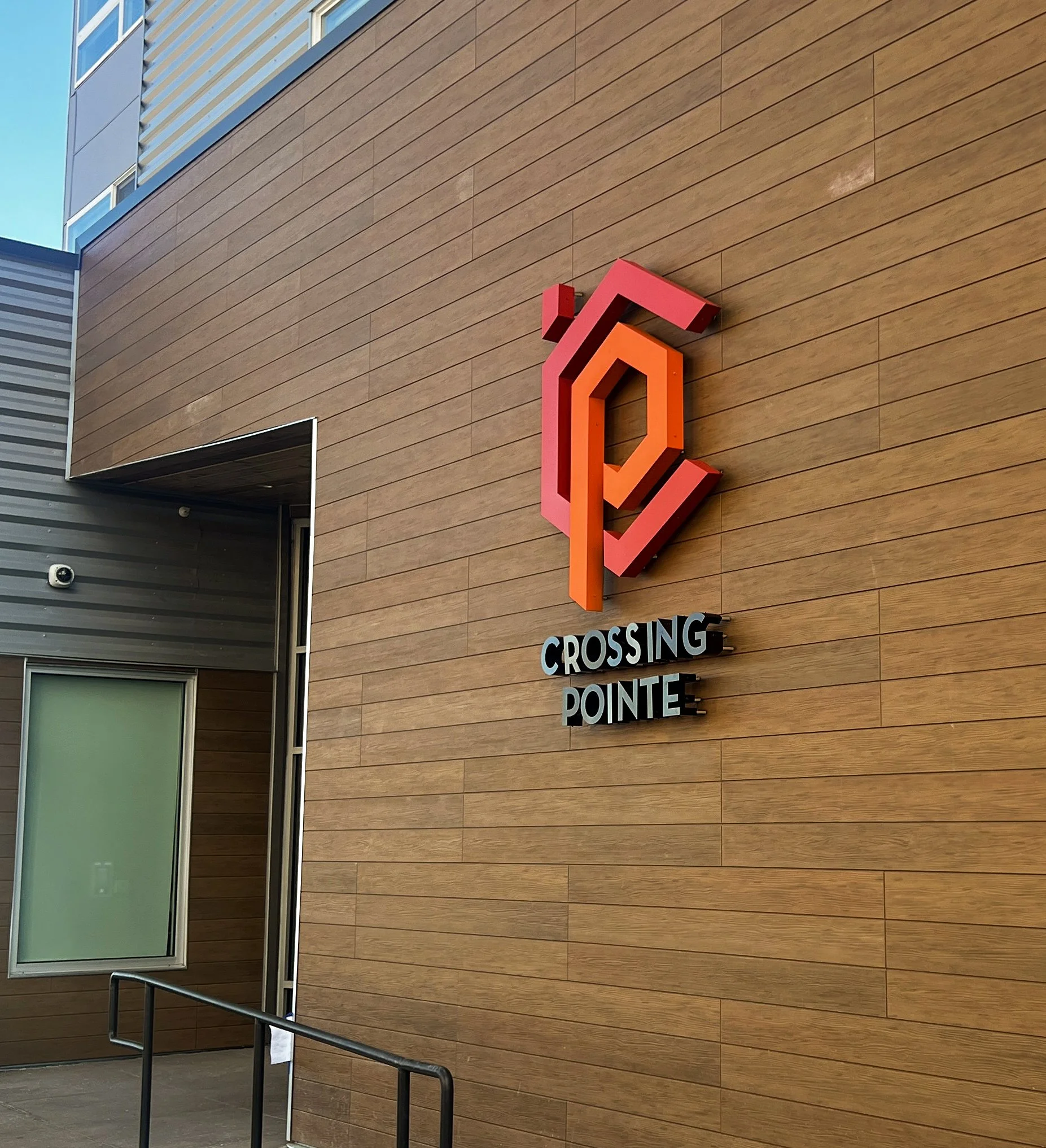 The image shows a large lit building sign on a modern building wall with brown and gray siding. A large, colorful sign is mounted on the wall with the words 'Crossing Pointe' and a geometric logo above it, featuring red and orange 3D shapes.