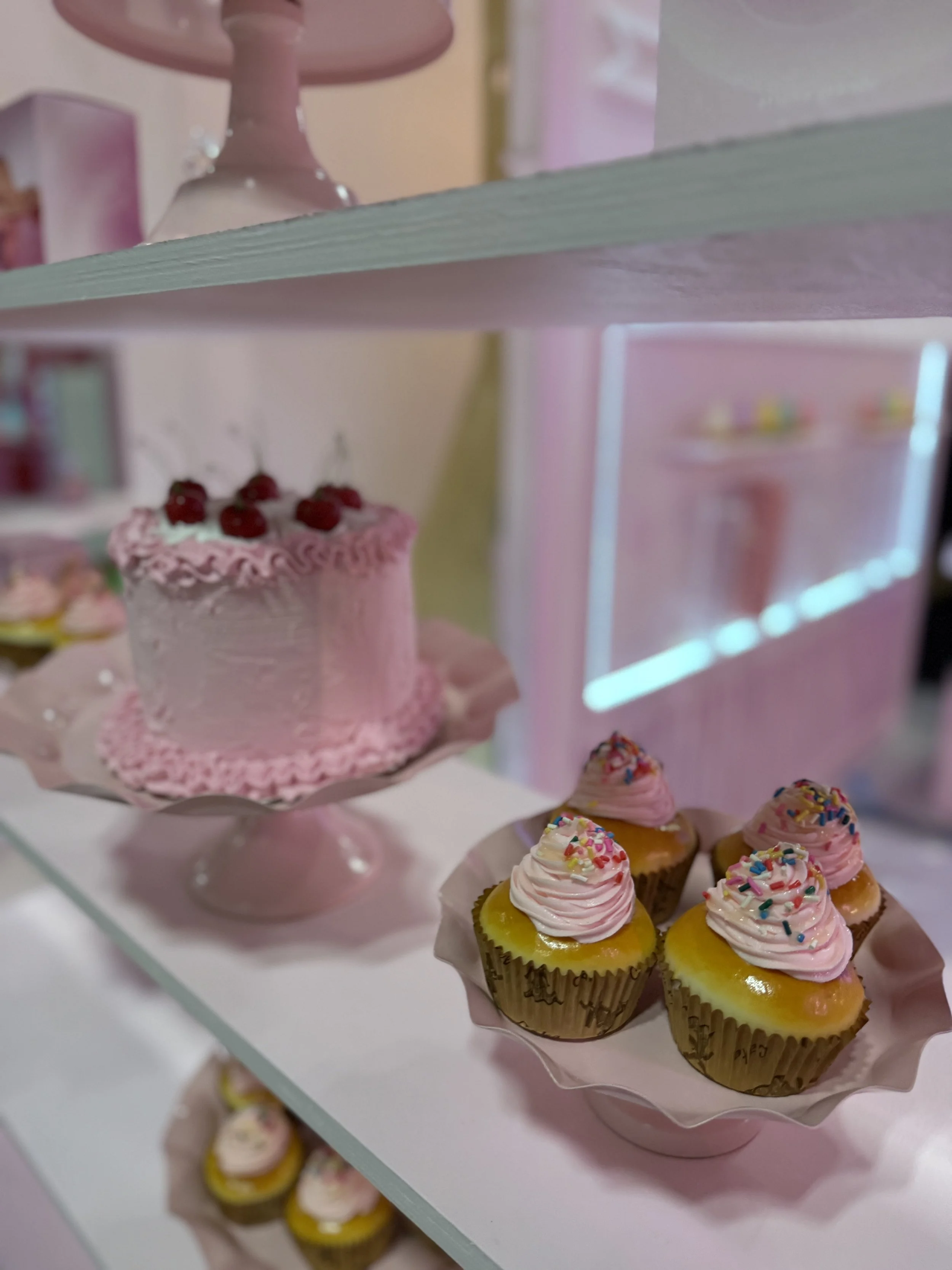 Display of pink frosted cake topped with cherries and mini cupcakes with pink frosting and rainbow sprinkles on a pink pedestal and white shelf.