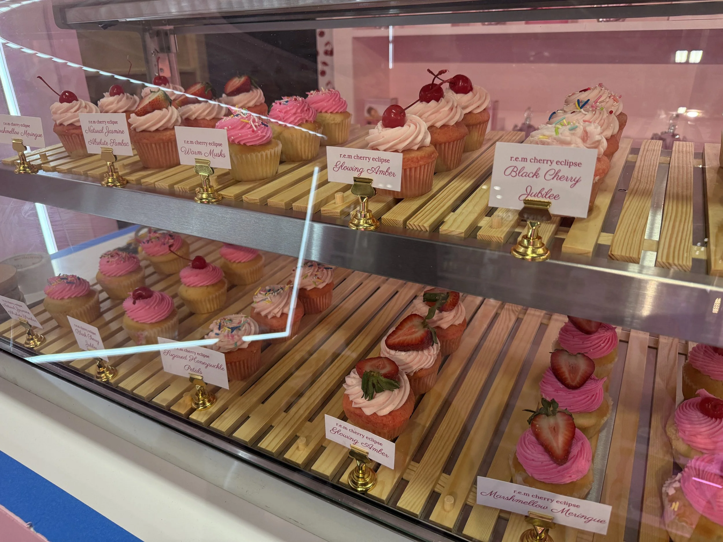 Display case filled with various decorated cupcakes, including pink and white frosting topped with cherries and strawberries, with small labels identifying each flavor.