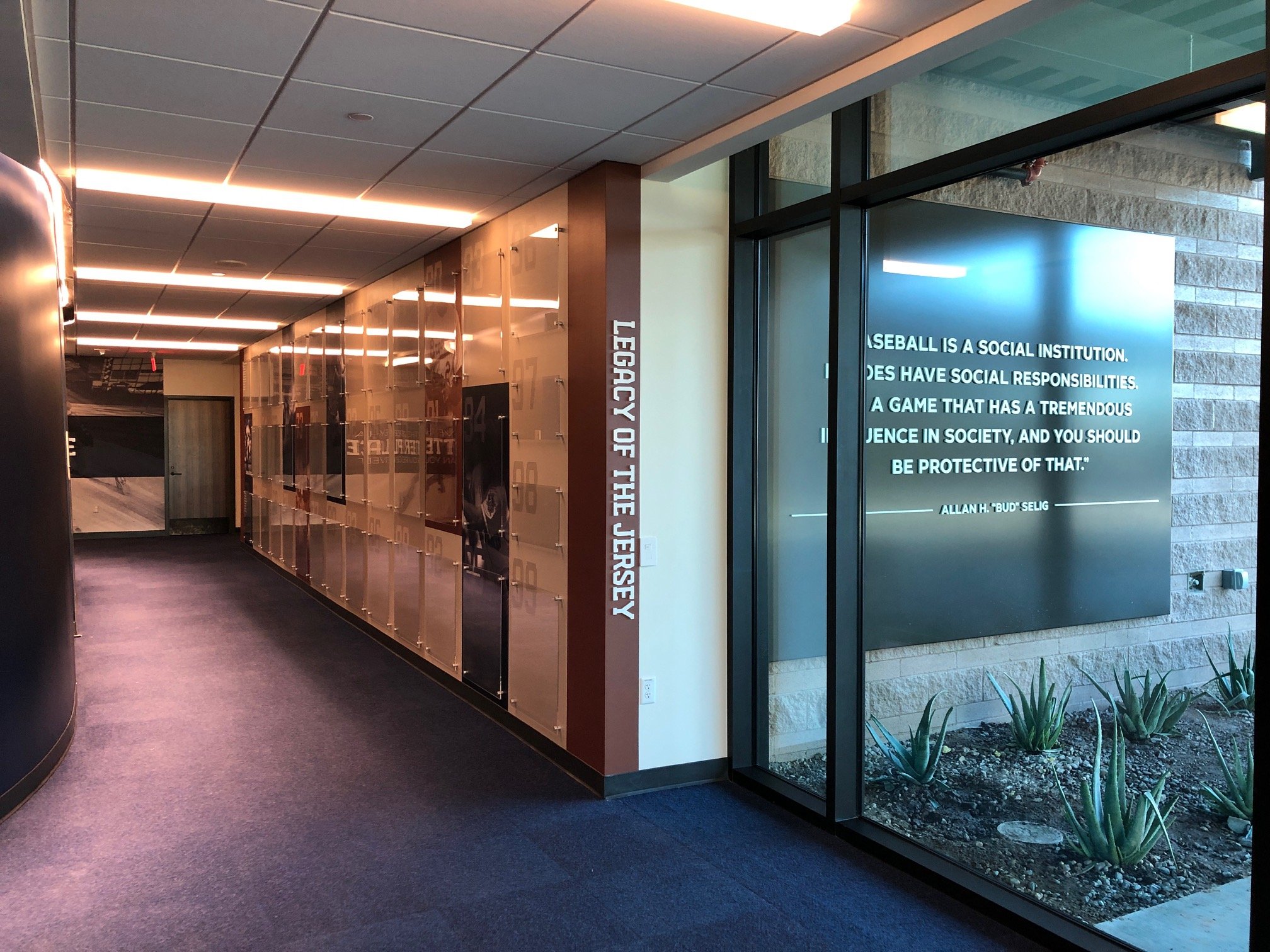 Indoor hallway with purple carpet, a wall of lockers, and a large window displaying a signage quote about baseball by Allan H. Selig, on a black background with white text for the MLB Milwaukee Brewers. 