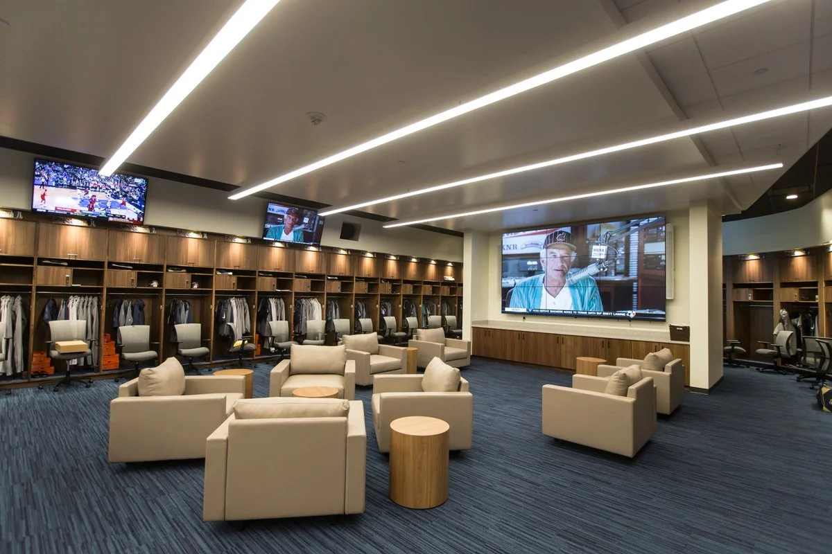 Luxury MLB baseball locker room for the Milwaukee Brewers Spring Training facility with beige seating area, multiple large screens on the wall, and lockers with clothing and gear inside.