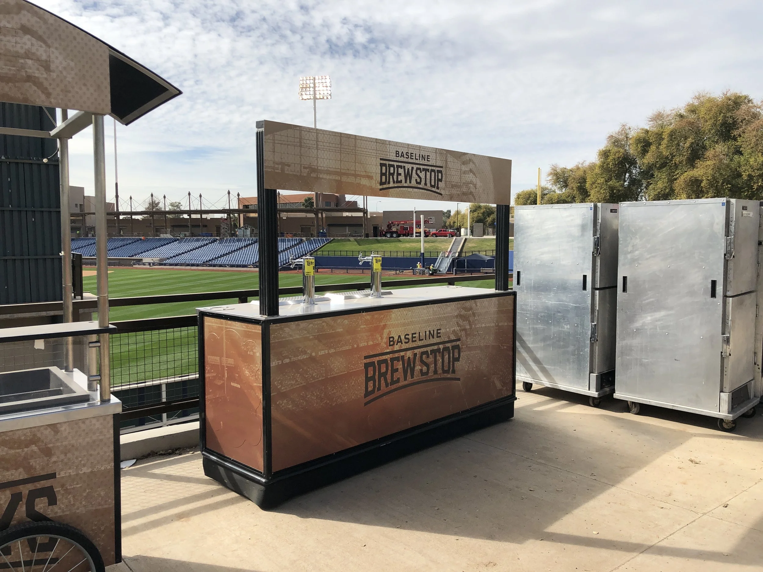 Outdoor beer stand labeled 'Baseline Brewstop' at a sports stadium, with football field and seating in the background.