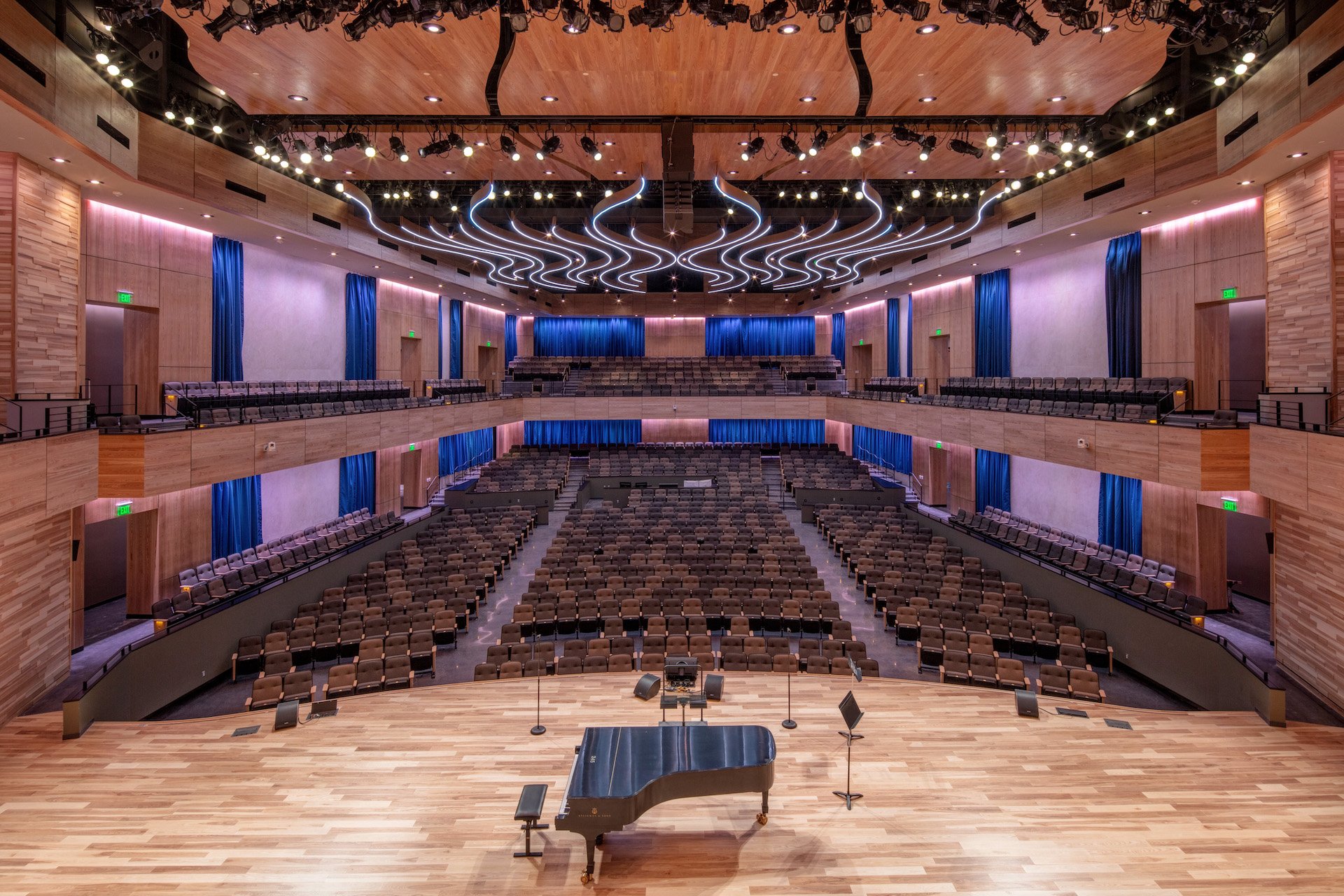 Empty concert hall with a grand piano on stage, surrounded by microphones and music stands, and rows of seating in the auditorium and balcony levels.