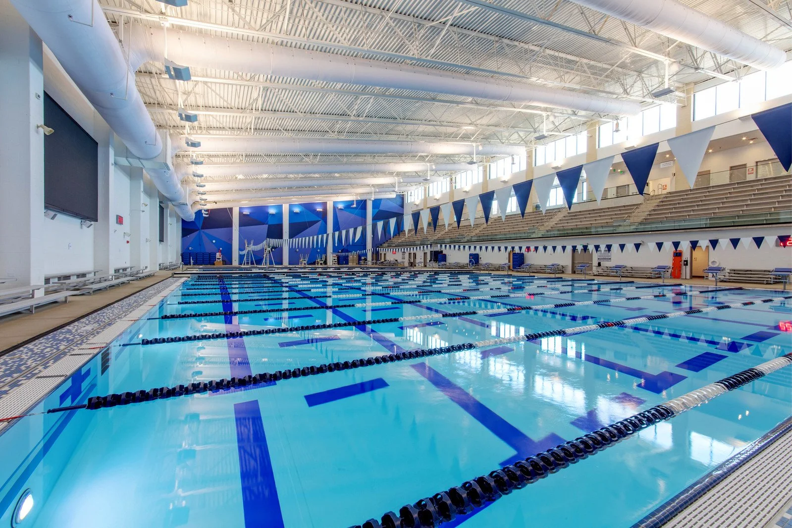 Indoor swimming pool with multiple lanes, blue and white triangular flags, large windows, and high ceiling.