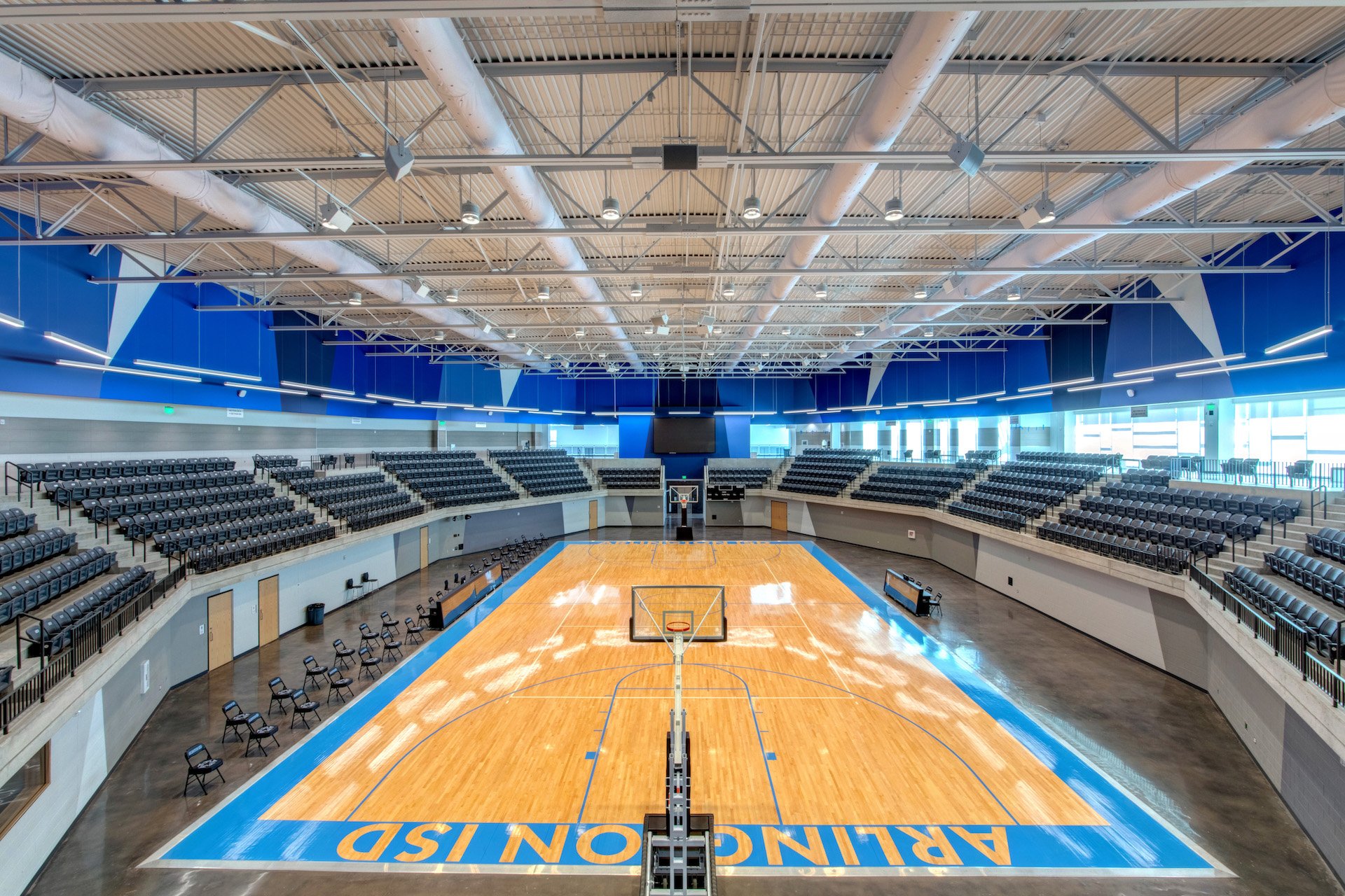 Arlington Sports Complex indoor basketball court with wooden flooring, black chairs along the sidelines, and blue graphics on walls with a large digital screen at the back. The court has the words "ARRINGTON ISD" painted at one end.